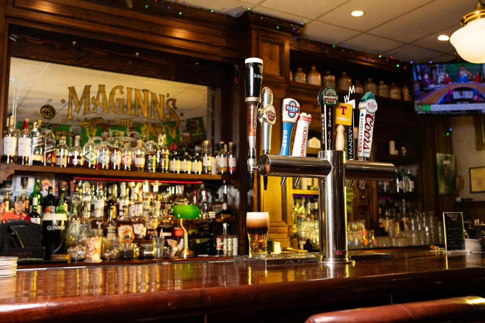 Interior of a pub with a wooden bar counter, several beer tap handles, and bottles of liquor in the background. A pint of beer with foam is on the counter.