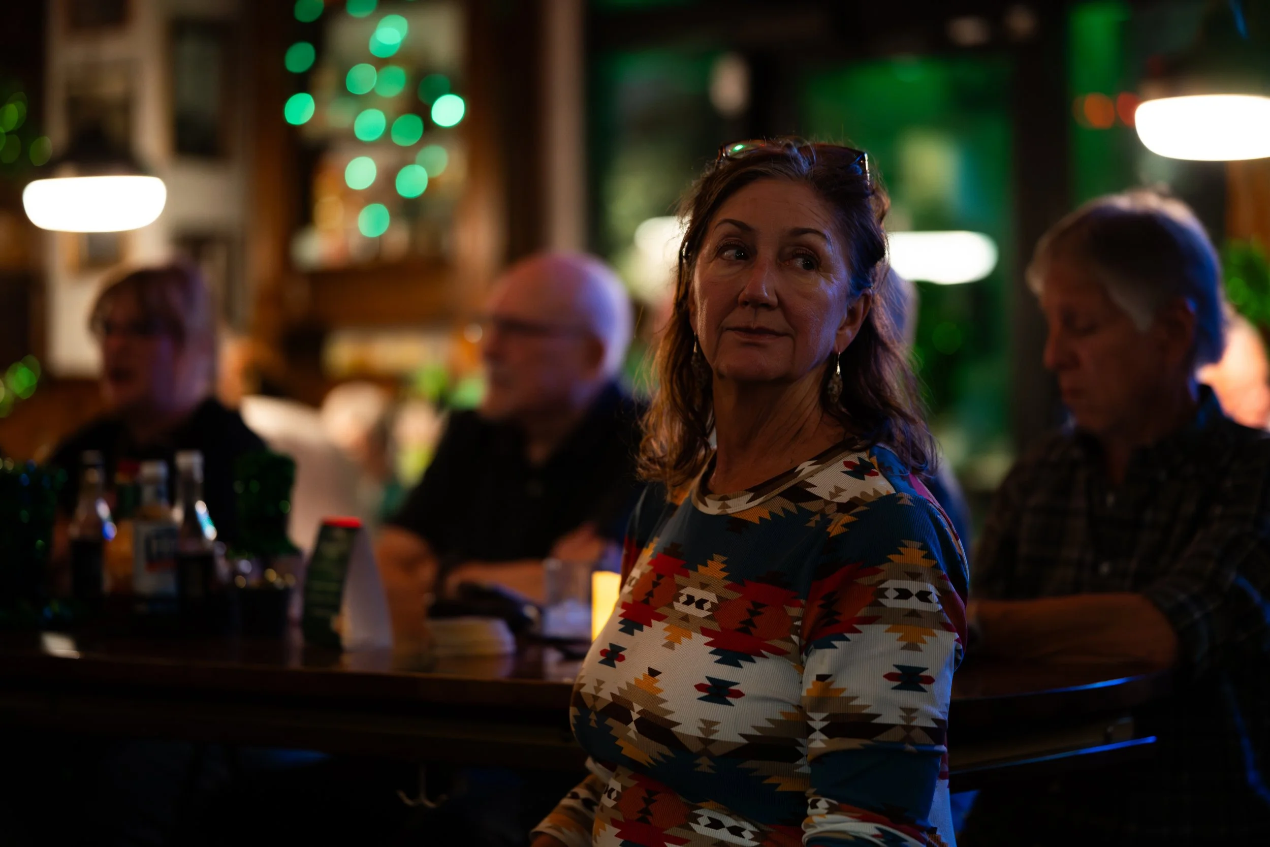 A woman in a patterned shirt looking to the side in a dimly lit bar or restaurant, with blurred figures of people sitting at a table behind her.