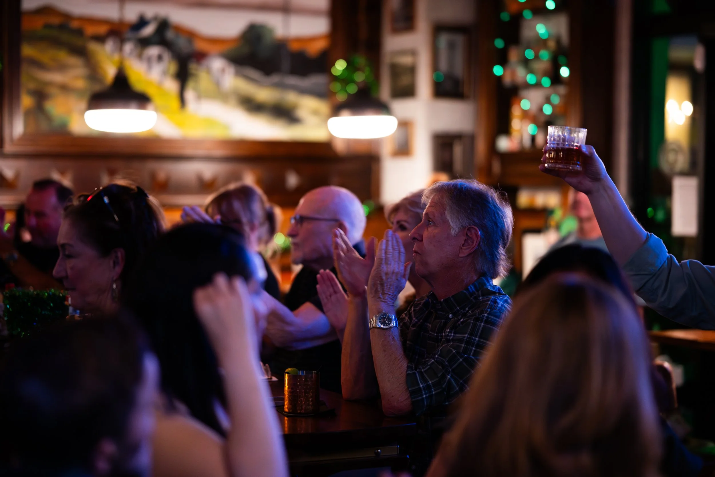People sitting in a dimly lit bar or restaurant, facing a person on stage, with someone raising a glass in the background