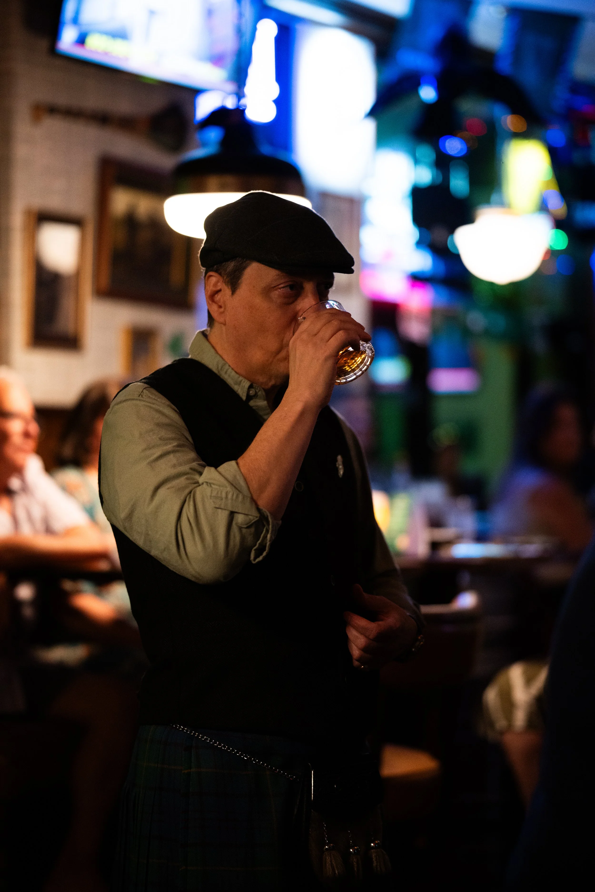 A man wearing a black beret and gray shirt, drinking a glass of beer inside a dimly lit pub or bar with colorful neon lights.