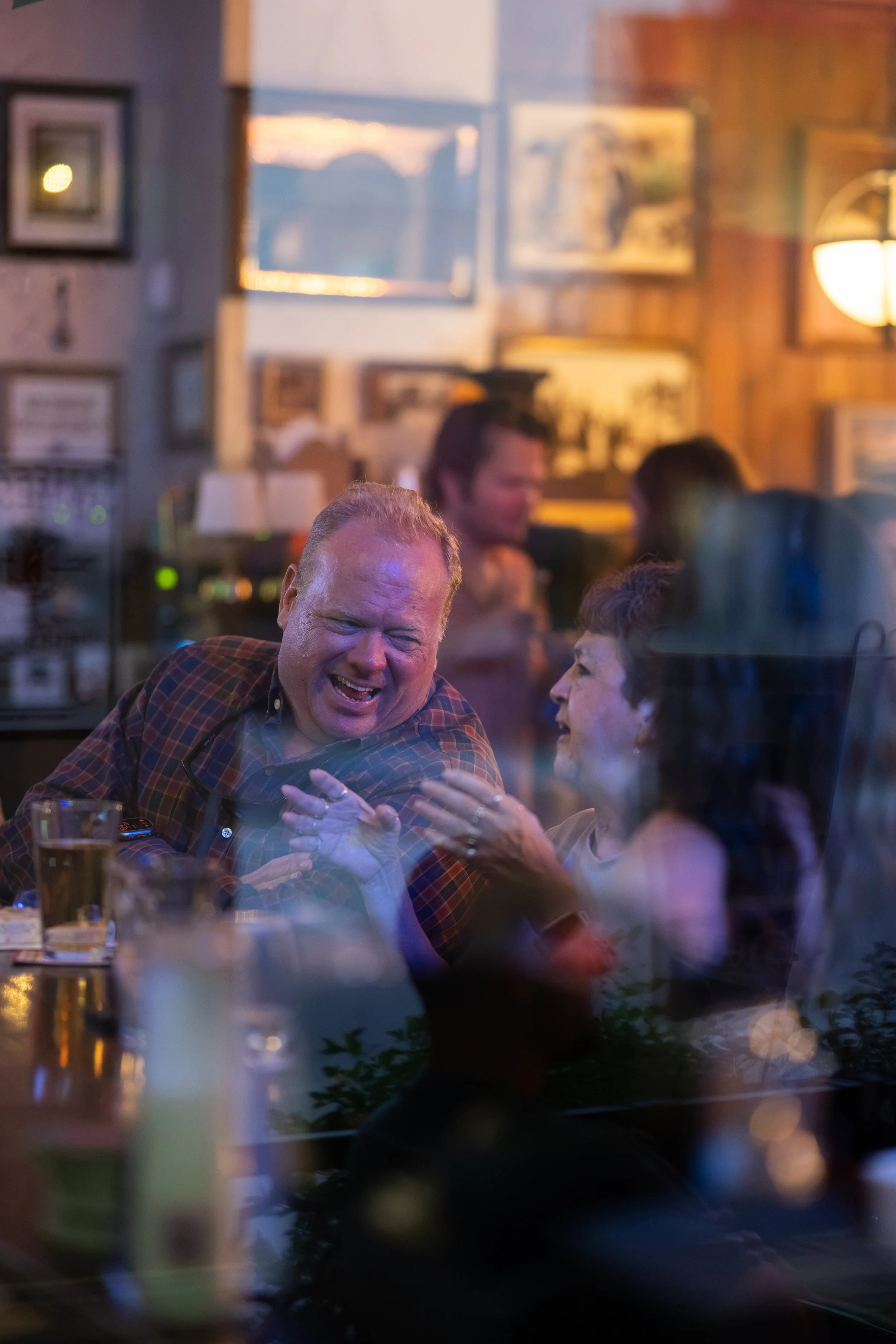 A group of people socializing and laughing in a cozy, warmly lit restaurant or pub. The scene includes a man in a plaid shirt smiling and engaging with an older woman, with blurred background details of framed pictures and warm lighting.