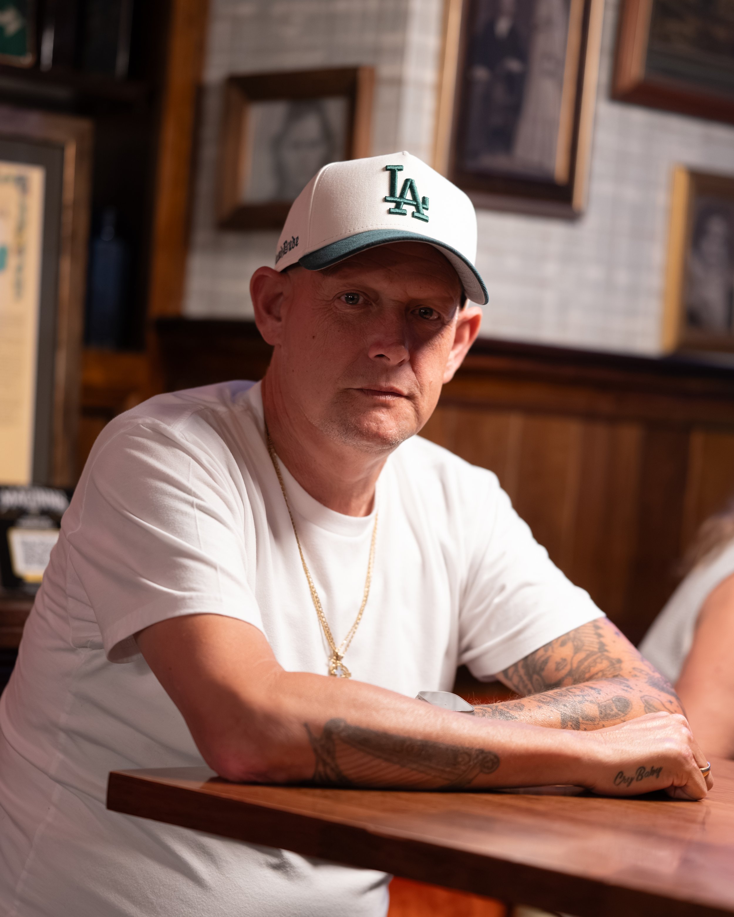 A man wearing a white Los Angeles Dodgers baseball cap, a white t-shirt, and a gold chain, sitting at a bar or restaurant with framed pictures on the wall behind him.
