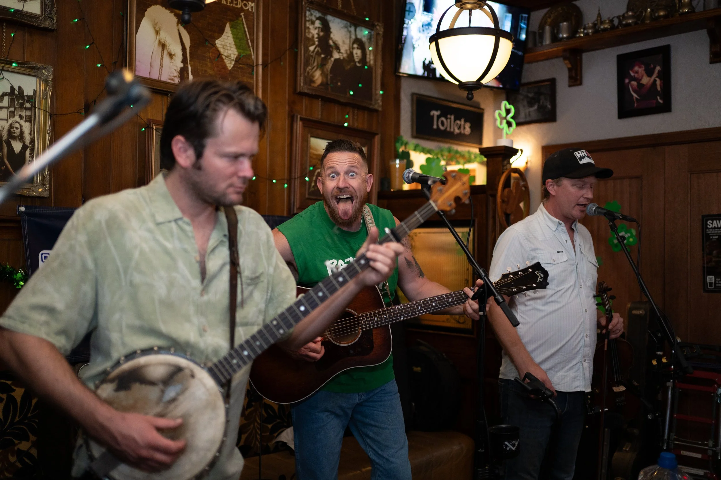 Three men performing music at a pub with wooden walls, Irish decorations, and framed photos, one playing a drum, one singing and playing guitar, and the third singing and playing a shaker.