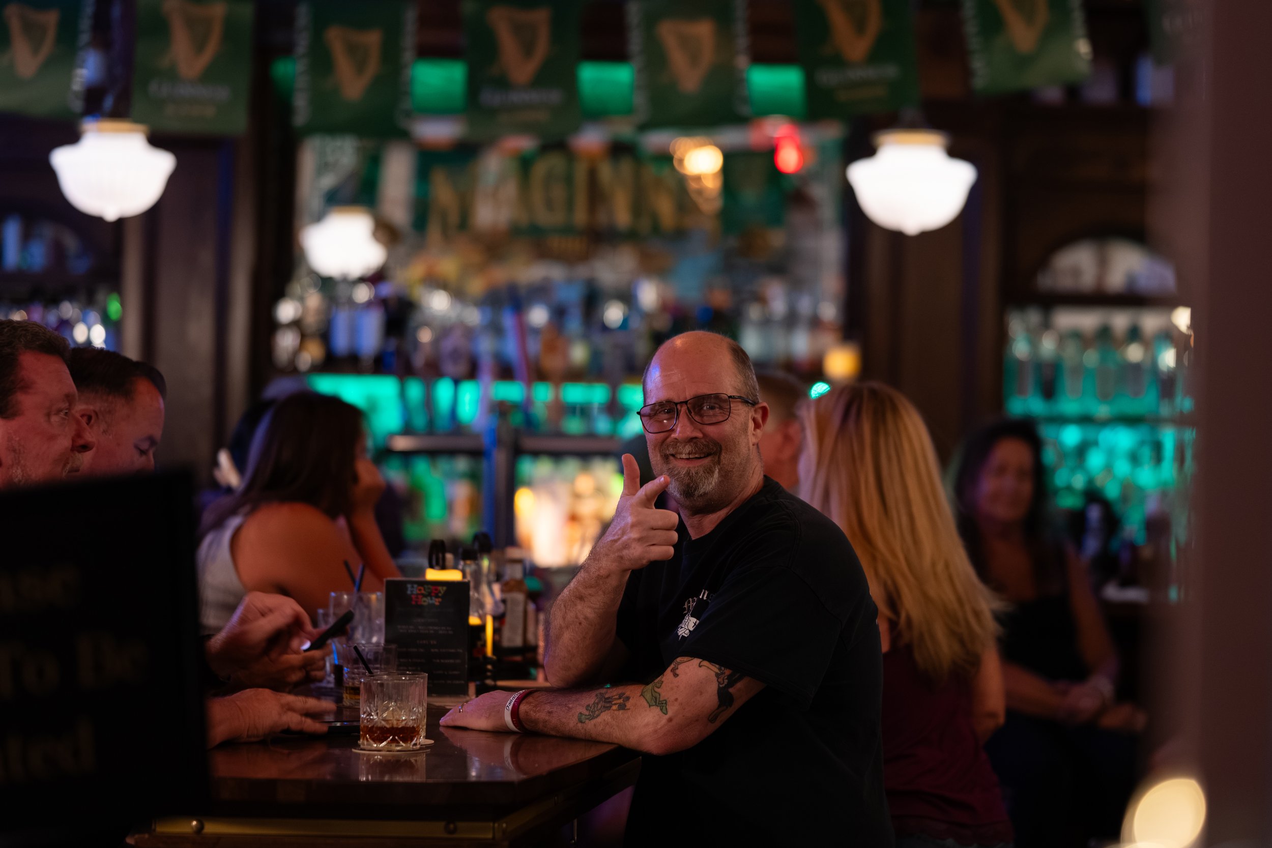 A man with tattoos and glasses smiling and giving a thumbs-up at a bar or pub, surrounded by other people, with warm and colorful lighting in the background.