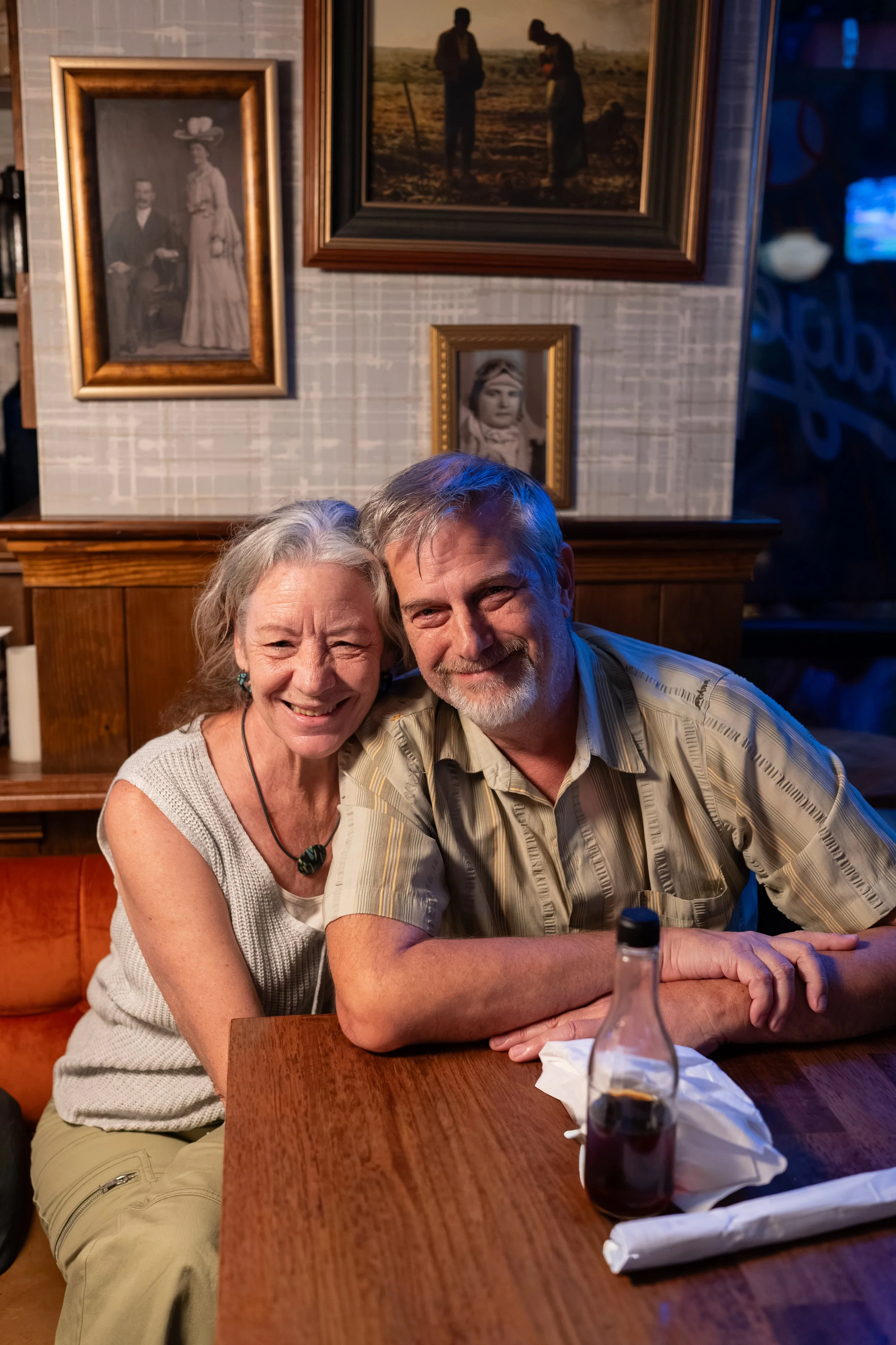 An elderly woman and a middle-aged man are sitting together at a wooden table in a cozy restaurant. They are smiling and appear happy. In the background, there are framed black-and-white photos on the wall. A bottle of dark soda and some napkins are 