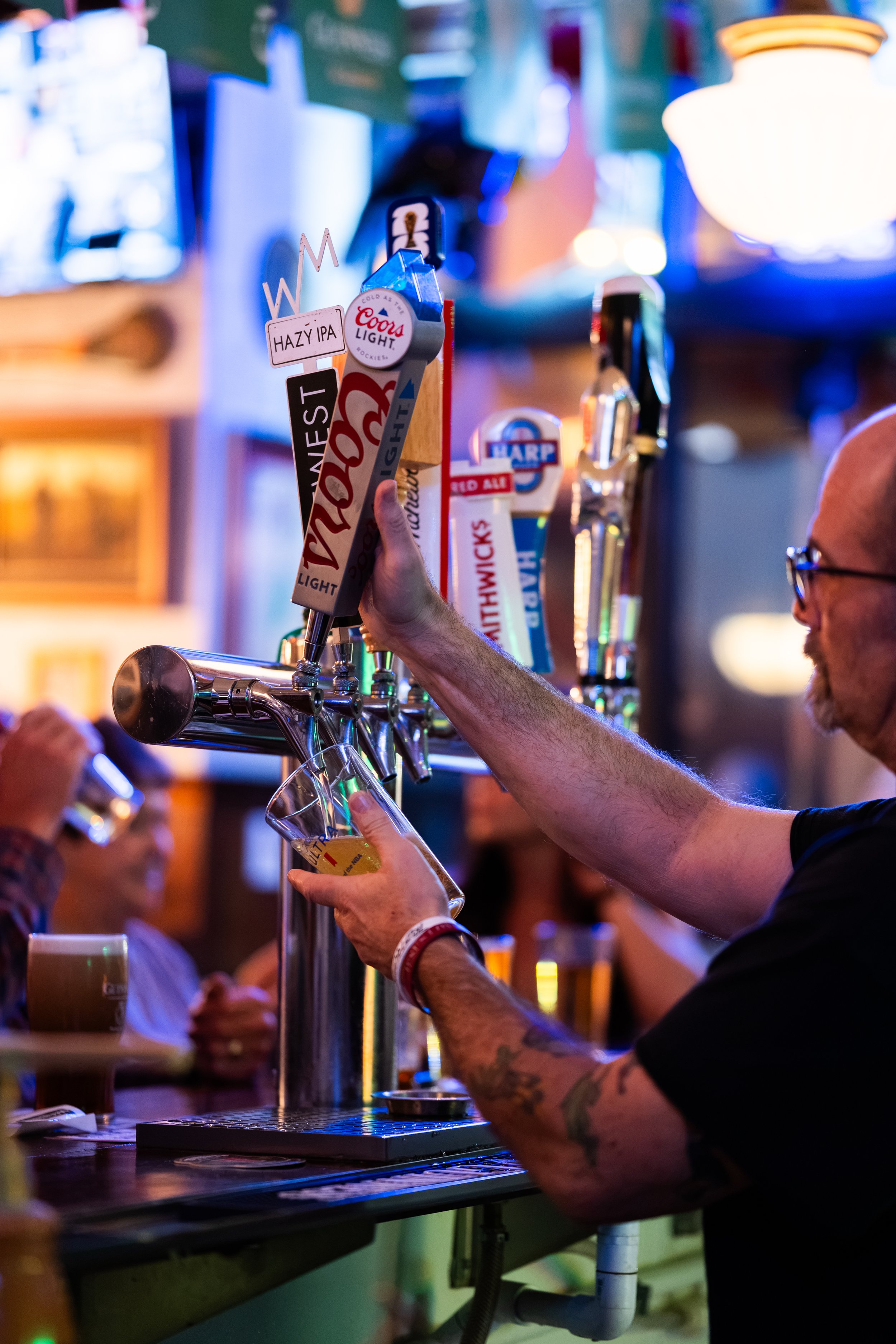 A person fills a glass with beer from a tap at a bar. The bar has multiple beer taps with different labels. The scene is illuminated with colorful lights, and other patrons are visible in the background.