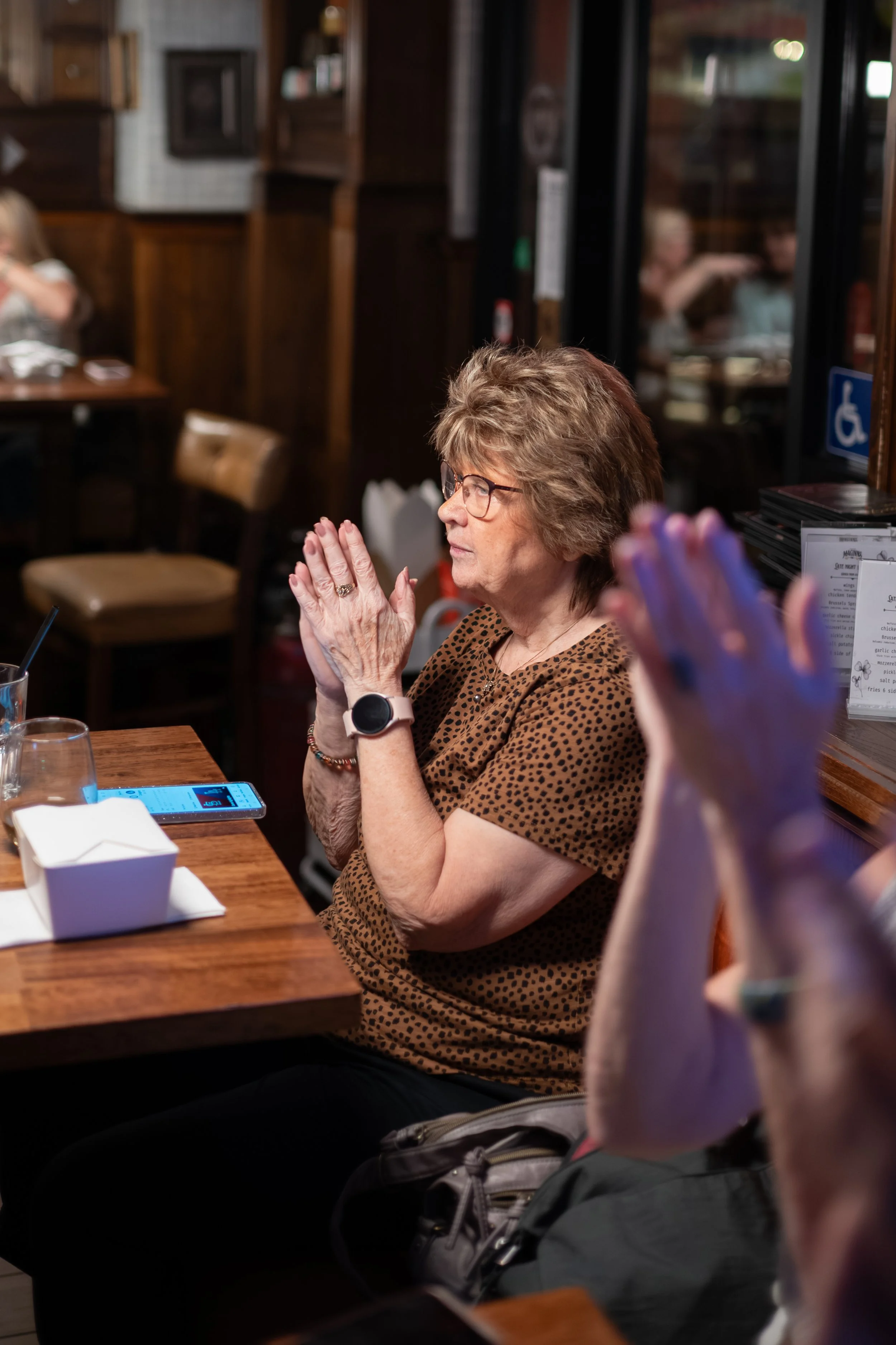 A woman with short, curly hair and glasses is sitting at a table, with her hands clasped together and eyes closed, appearing to be praying or meditating. She is wearing a leopard print top and has a smartwatch on her left wrist. In front of her is a 