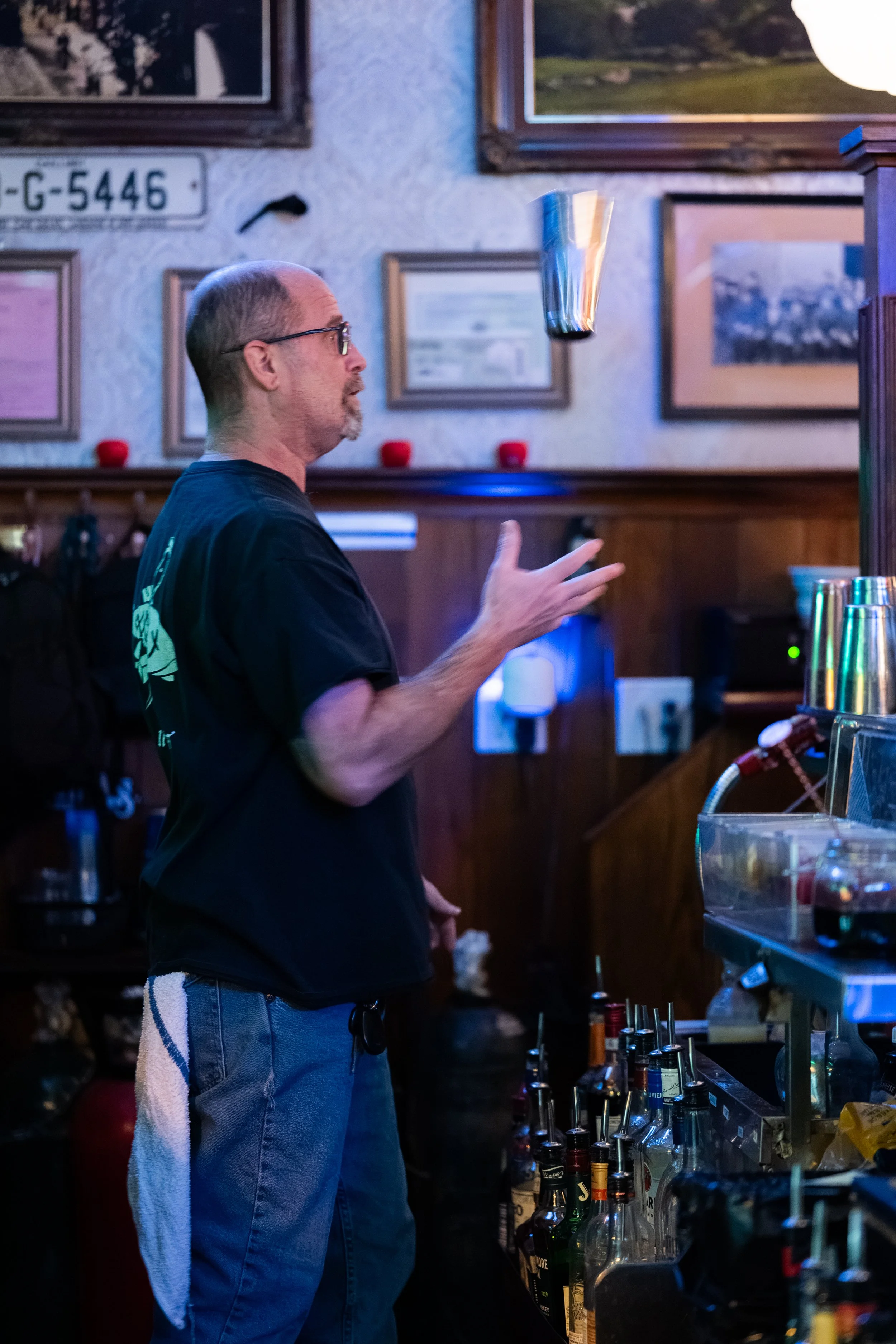 A man standing at a bar counter, tossing a cocktail shaker into the air. The bar has a variety of bottles, glasses, and bar tools. The background features framed pictures and a license plate on the wall.