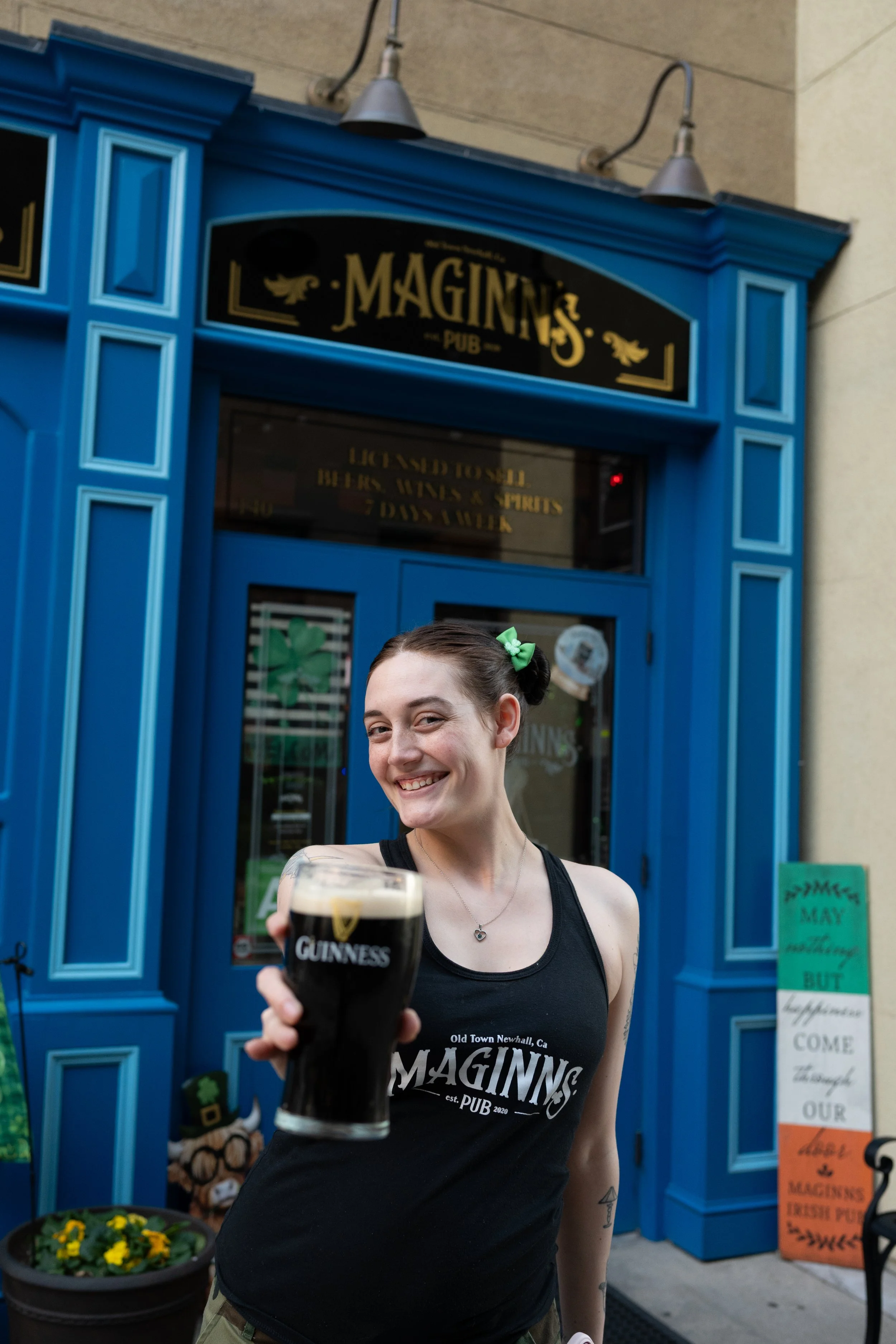 A woman smiling and holding a pint of Guinness beer in front of a pub named 'Maggins'.