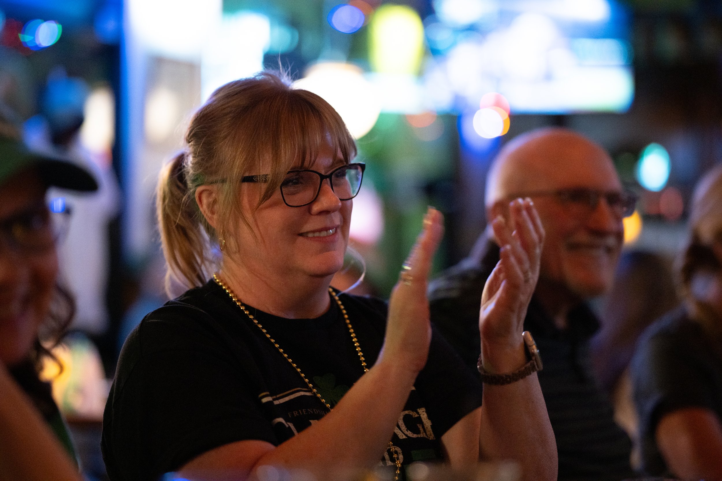 A group of people, including a woman with glasses and a man with glasses, are at a lively event, some clapping and smiling.