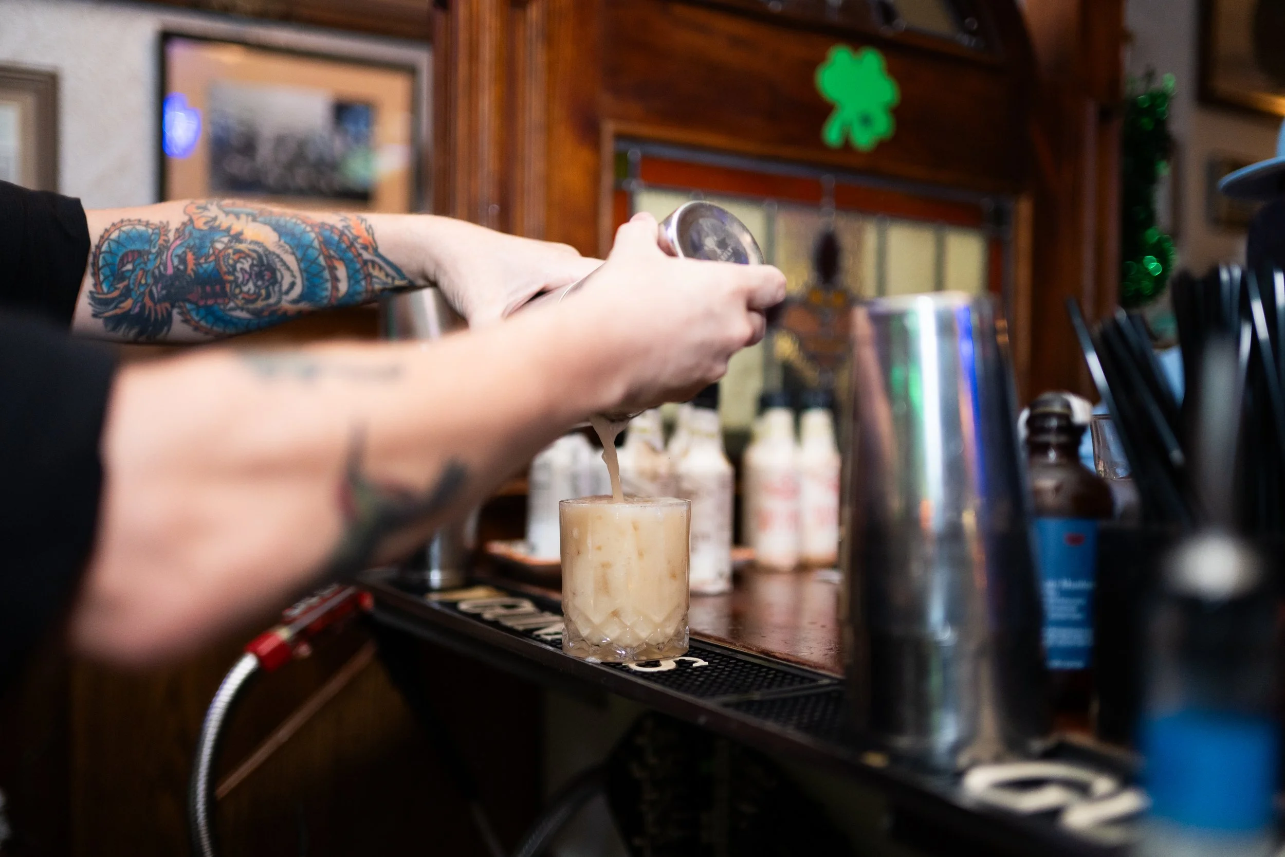 A bartender with tattoos on their arms pours a drink from a shaker into a glass at a bar, with bottles and bar tools visible in the background.