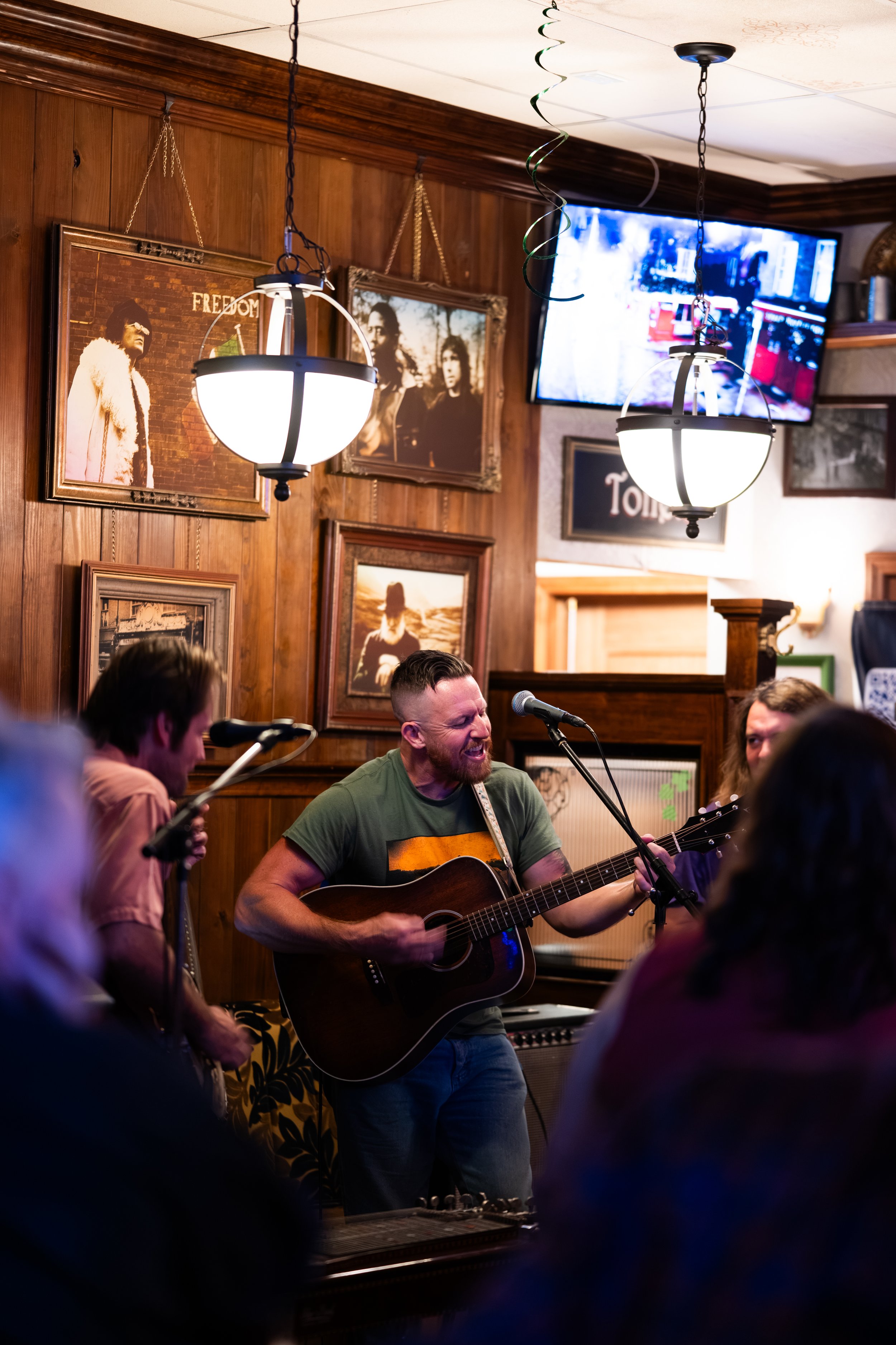 A man playing an acoustic guitar and singing into a microphone in a cozy, wood-paneled room with framed photos and a TV on the wall, surrounded by an audience.