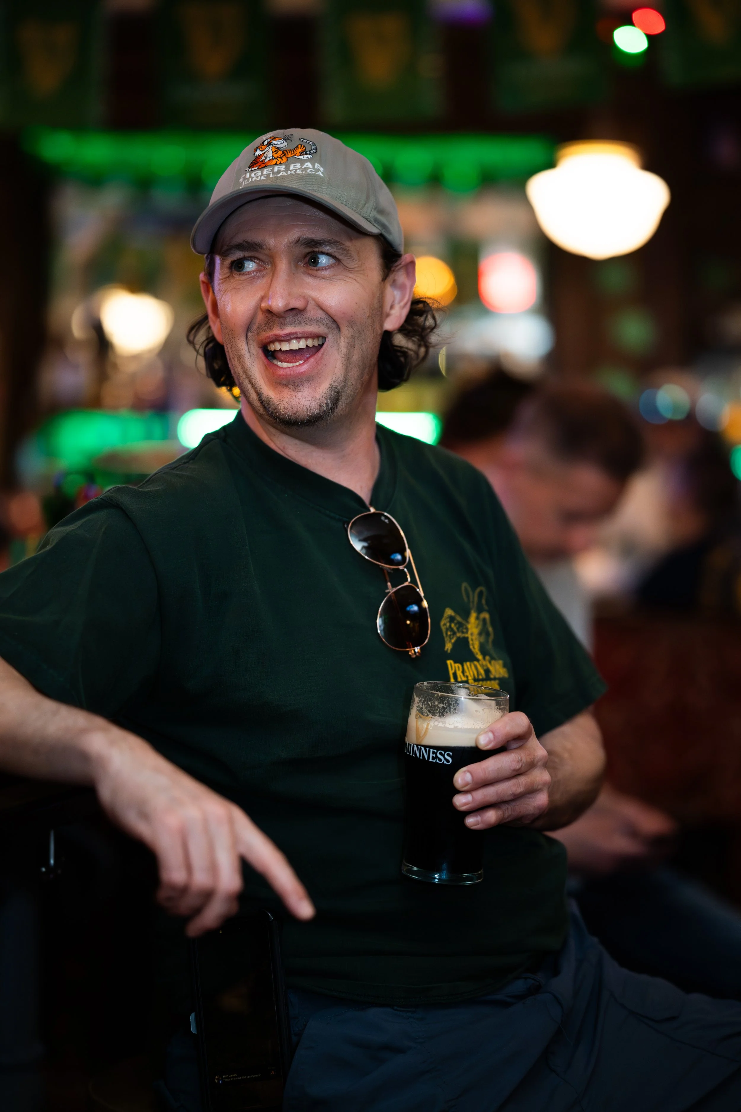 A man wearing a gray cap, sunglasses hanging on his shirt, and a black T-shirt with a yellow logo, holding a pint of Guinness beer, in a lively bar with colorful lights and blurred patrons in the background.