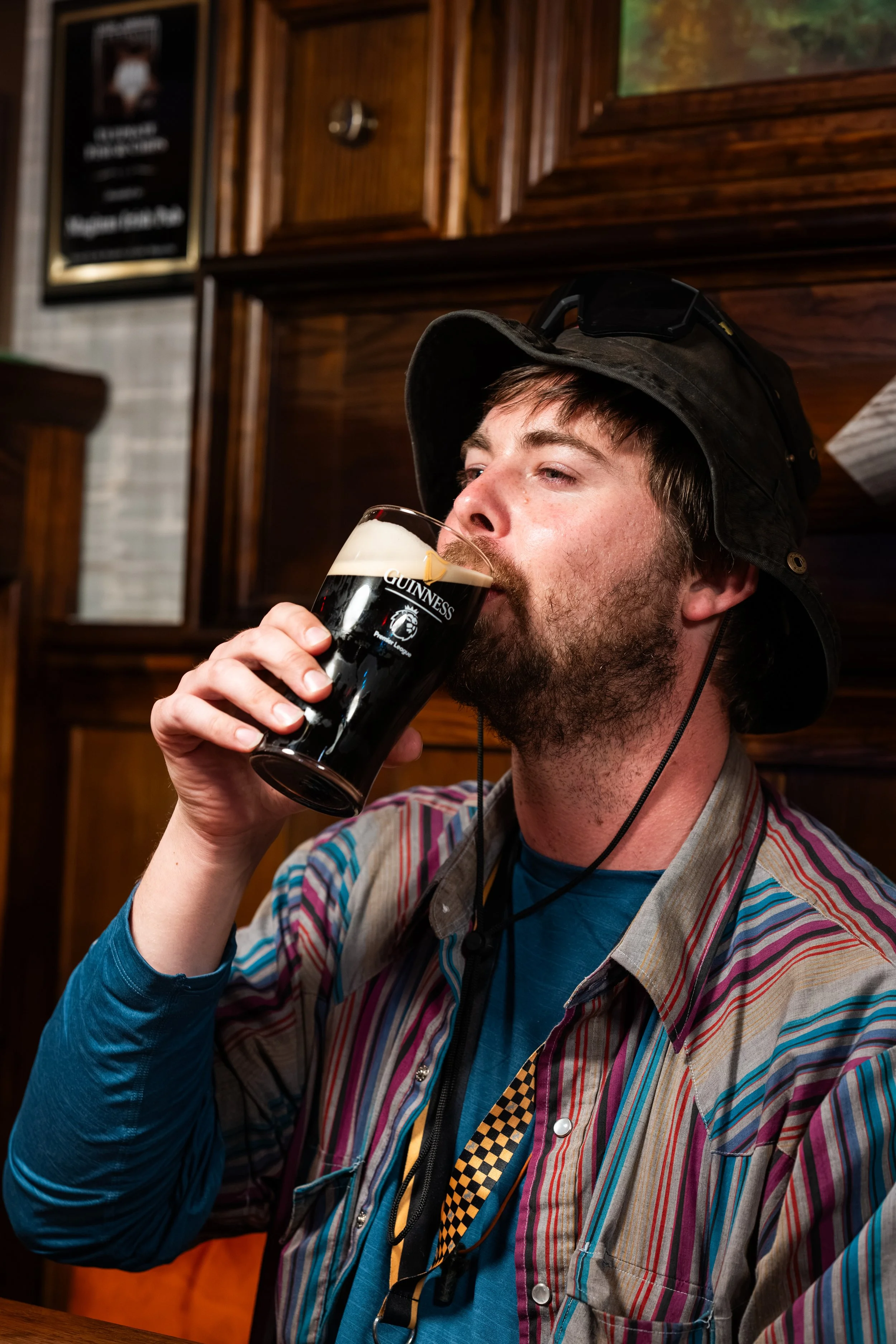 Man with a beard drinking Guinness beer in a pub, wearing a striped shirt, a hat, and sunglasses on his head.