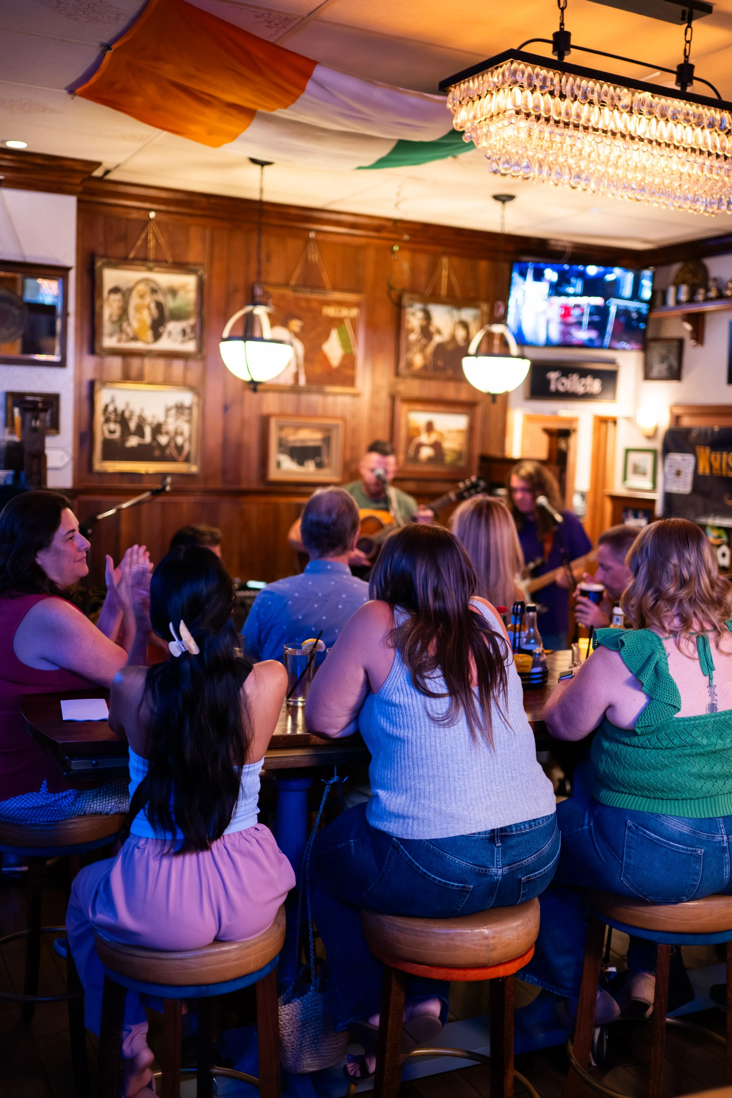 People gathered in a cozy pub listening to live music, with a man playing guitar and crowding around tables, framed pictures on wood-paneled walls, hanging lamps, and a television showing a sports game.