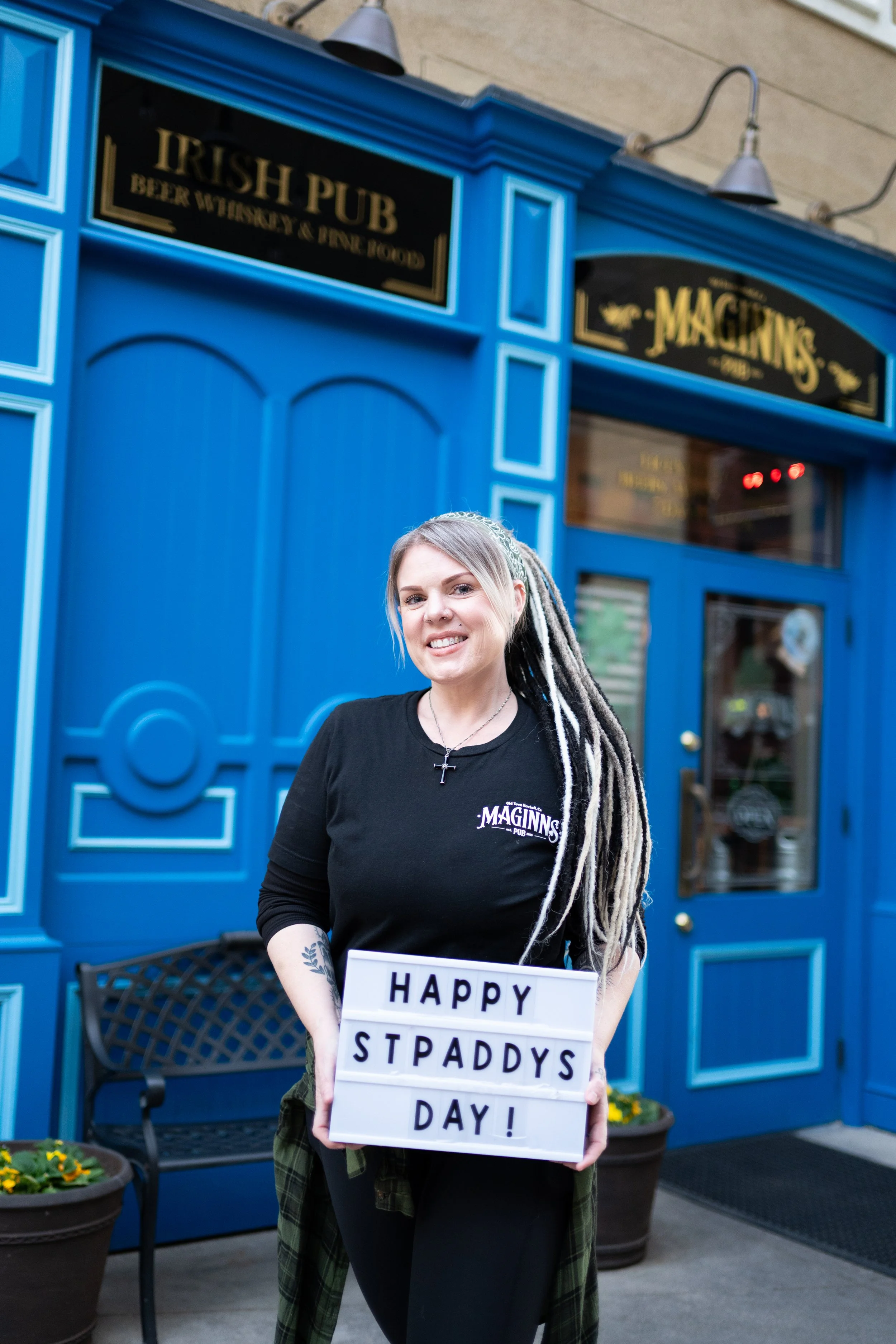 A woman with gray dreadlocks and a black shirt holding a sign that reads 'Happy St. Paddy's Day!' outside a blue Irish pub.