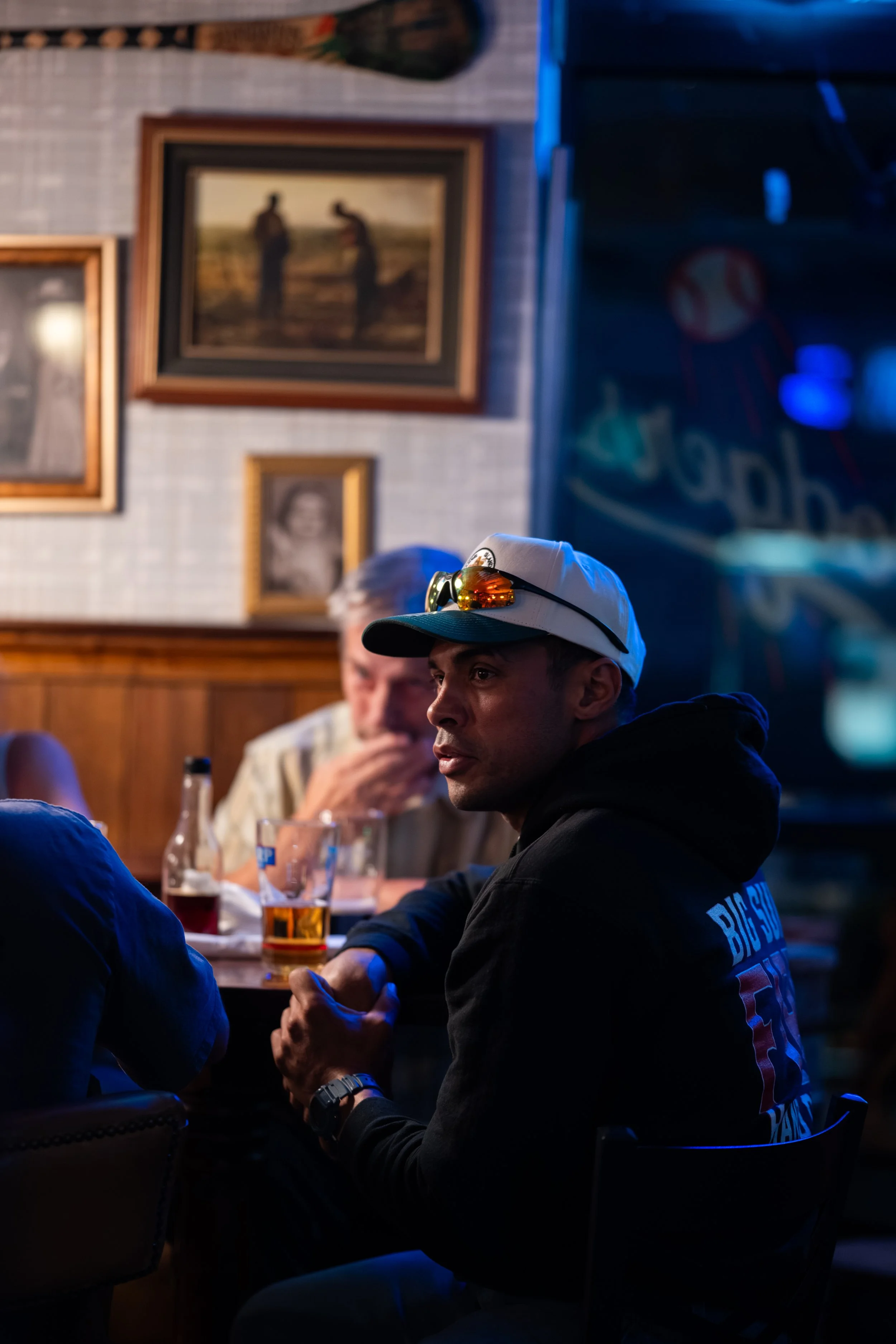 People gathered at a bar or restaurant, sitting around a table with drinks, with one man in the foreground wearing a white cap, black hoodie with 'BIG SUR' written on the back, and sunglasses on top of his cap, in a dimly lit setting with framed pict