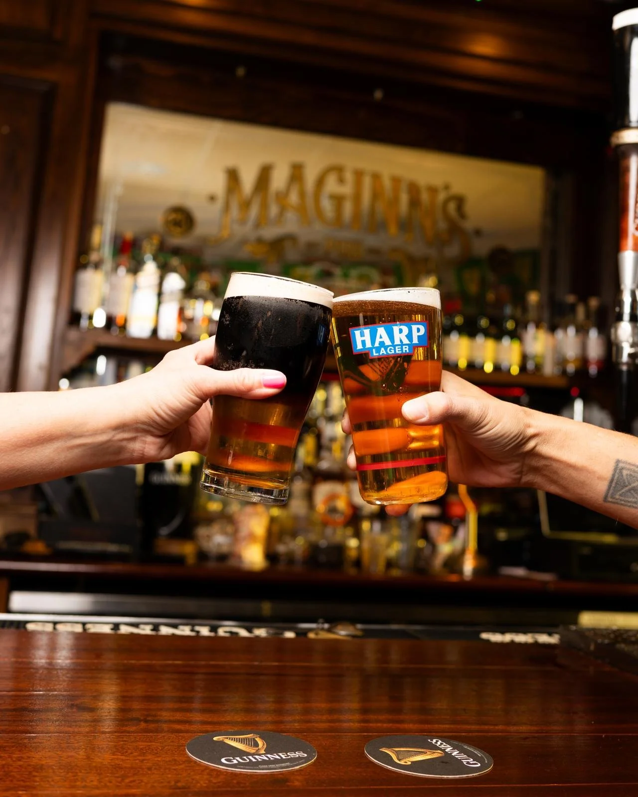 Two people clinking glasses of beer, one with dark stout and the other with HARP Lager, inside a bar with wooden decor and shelves of liquor in the background.