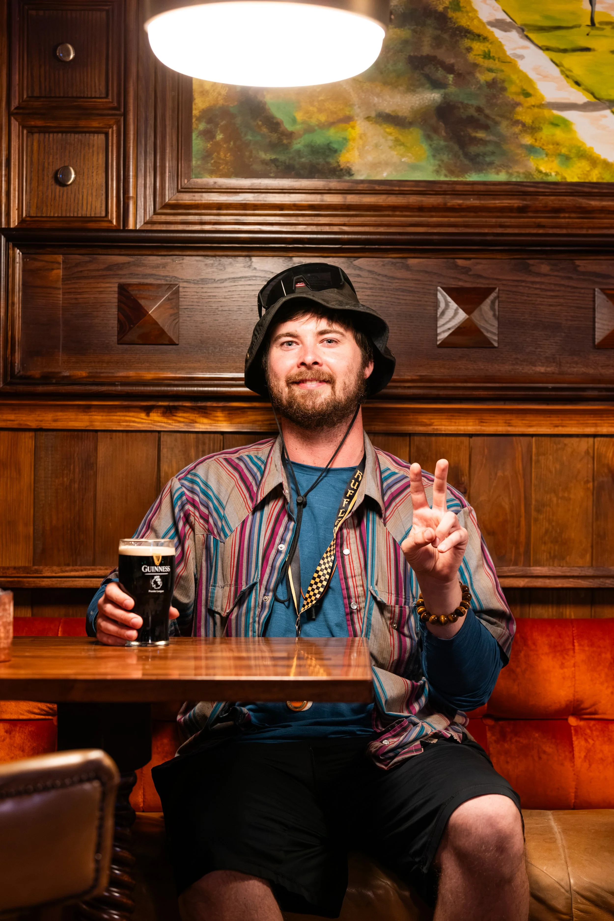 A man with a beard and brown hair, wearing a patterned shirt, black shorts, a bucket hat, and a beaded bracelet, sitting in a wooden booth at a restaurant. He is holding a glass of Guinness beer and smiling with a peace sign gesture. There is a paint