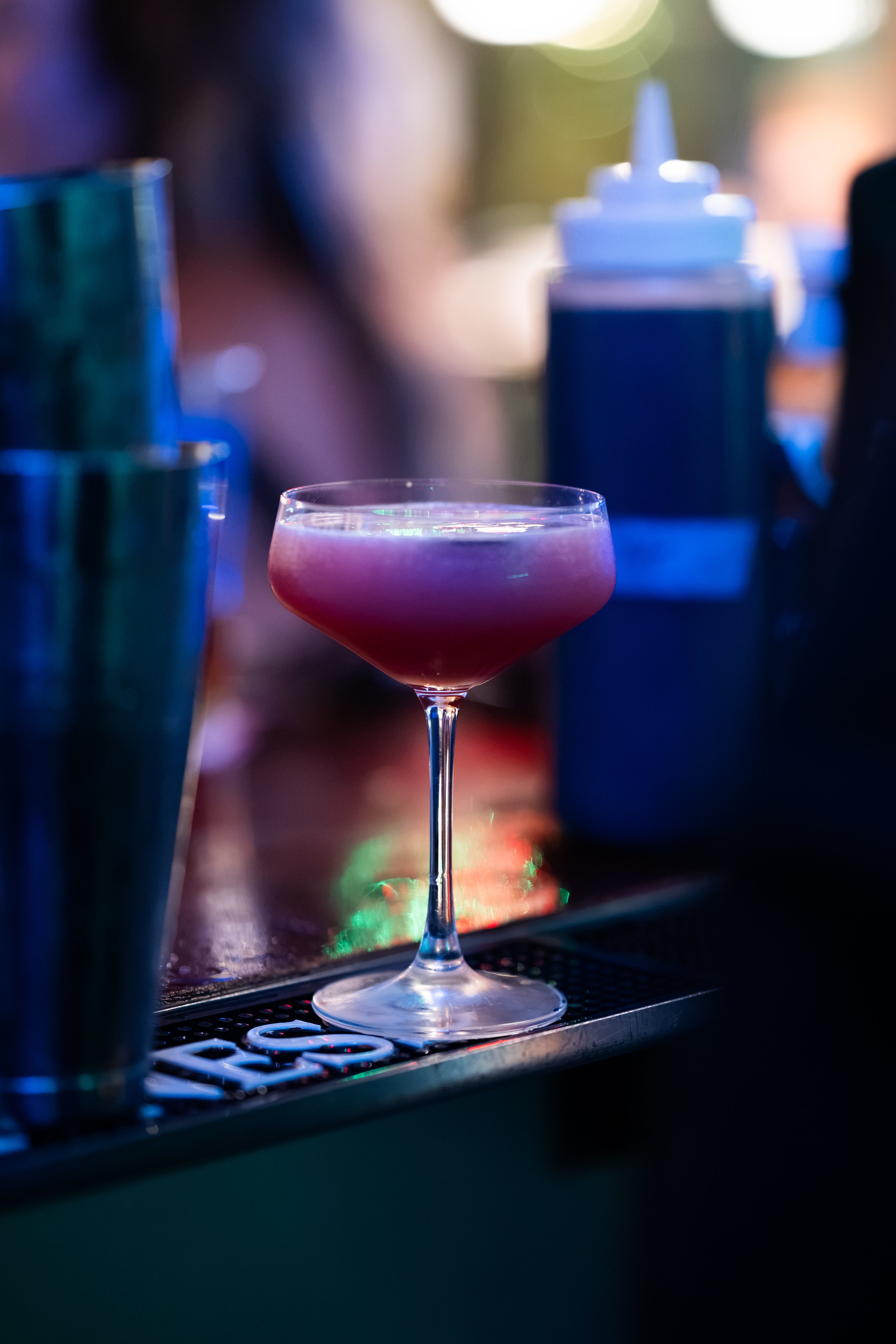A pink cocktail in a coupe glass on a bar counter with blurred bar tools and bottles in the background, illuminated by colorful lights.