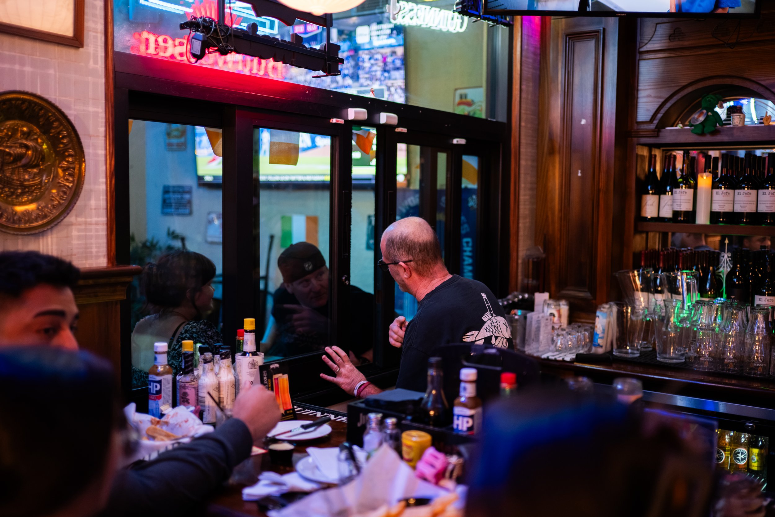 A group of people gathered around a bar counter inside a dimly lit restaurant or pub, engaging in a conversation. The bar is stocked with various bottles and bar equipment. Neon lights and a television screen displaying a sports game are visible in t