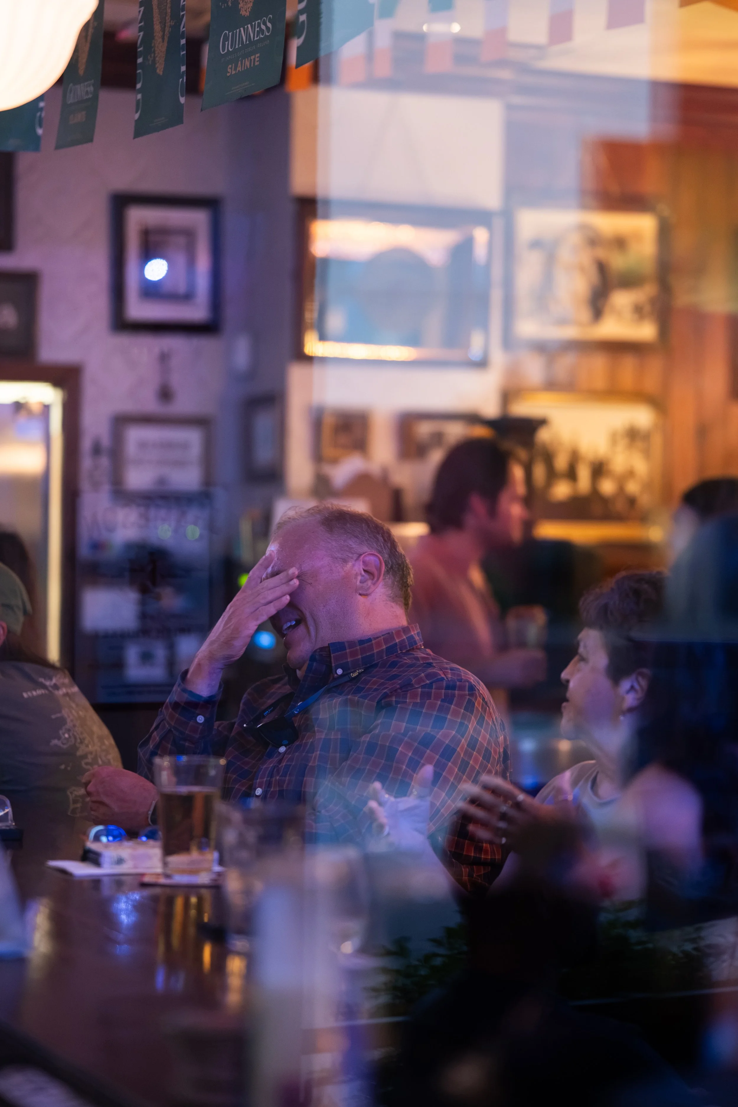 People sitting at a table in a pub or restaurant, engaging in conversation, with framed pictures on the wall and banners hanging from the ceiling.