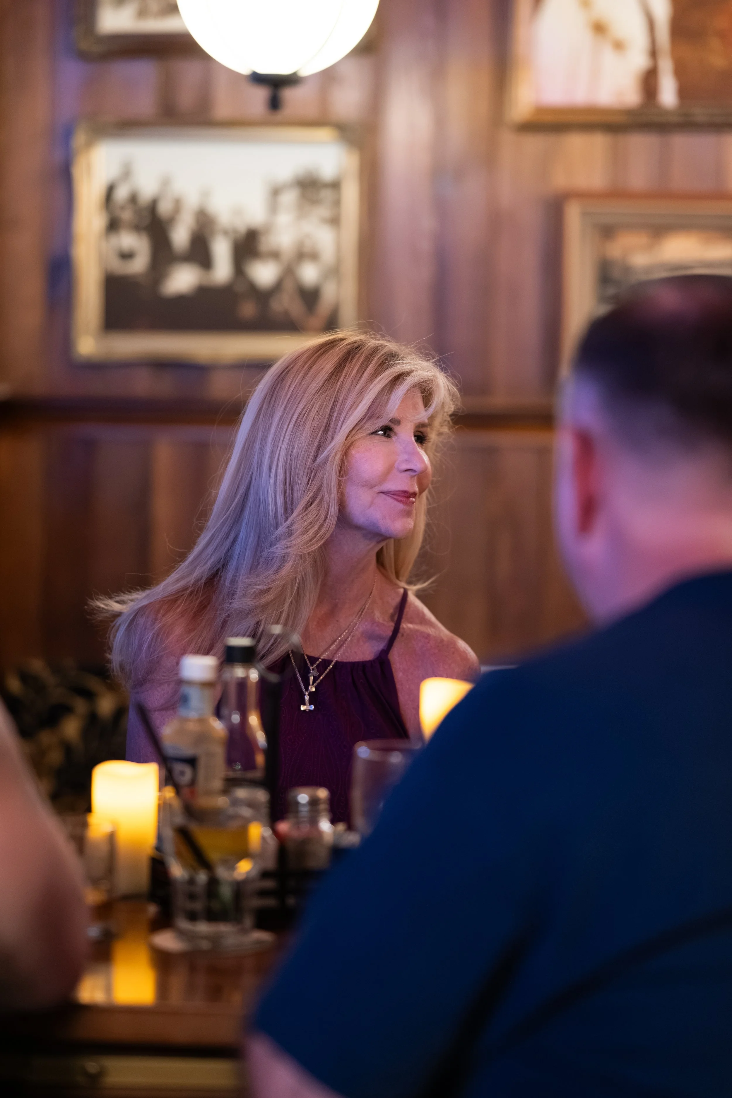 A woman with long blonde hair smiling in a dimly lit restaurant with wooden walls and framed pictures, sitting across from a man whose back is visible, surrounded by bottles and candlelight.