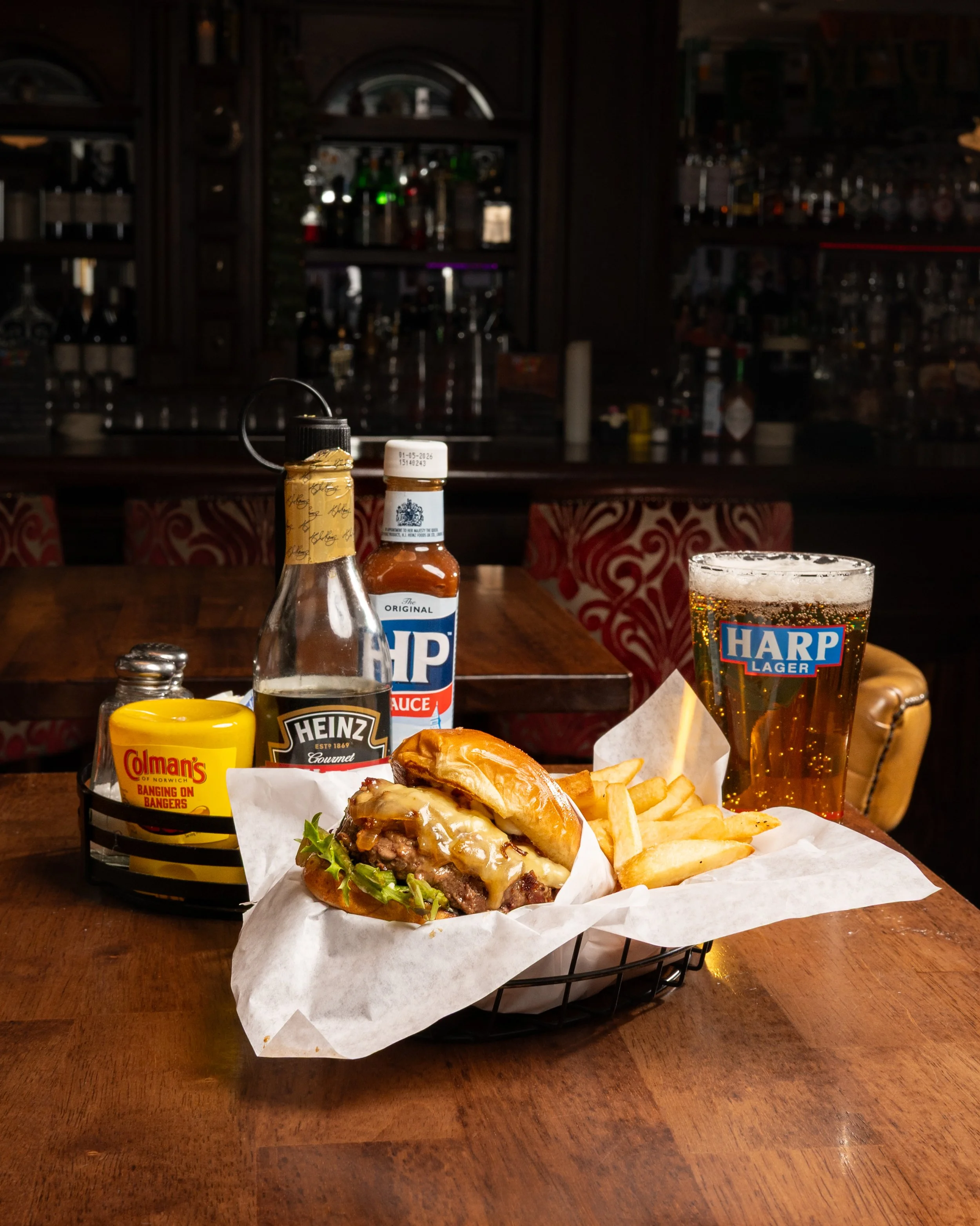 Cheeseburger with lettuce and cheese, French fries, a glass of beer, ketchup, mustard, and condiments on a wooden table in a bar or restaurant.