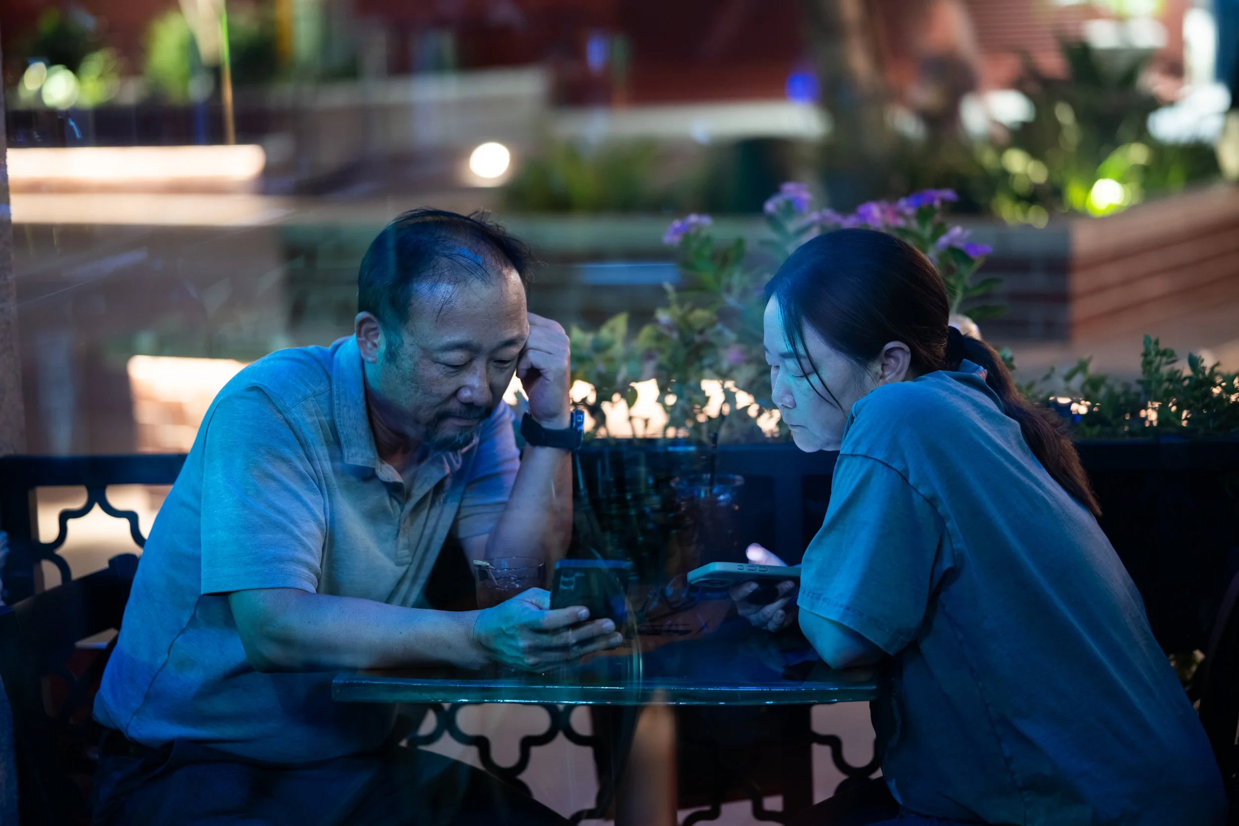 Two people sitting at a table indoors, looking at a mobile device, with vibrant lighting and plants in the background.