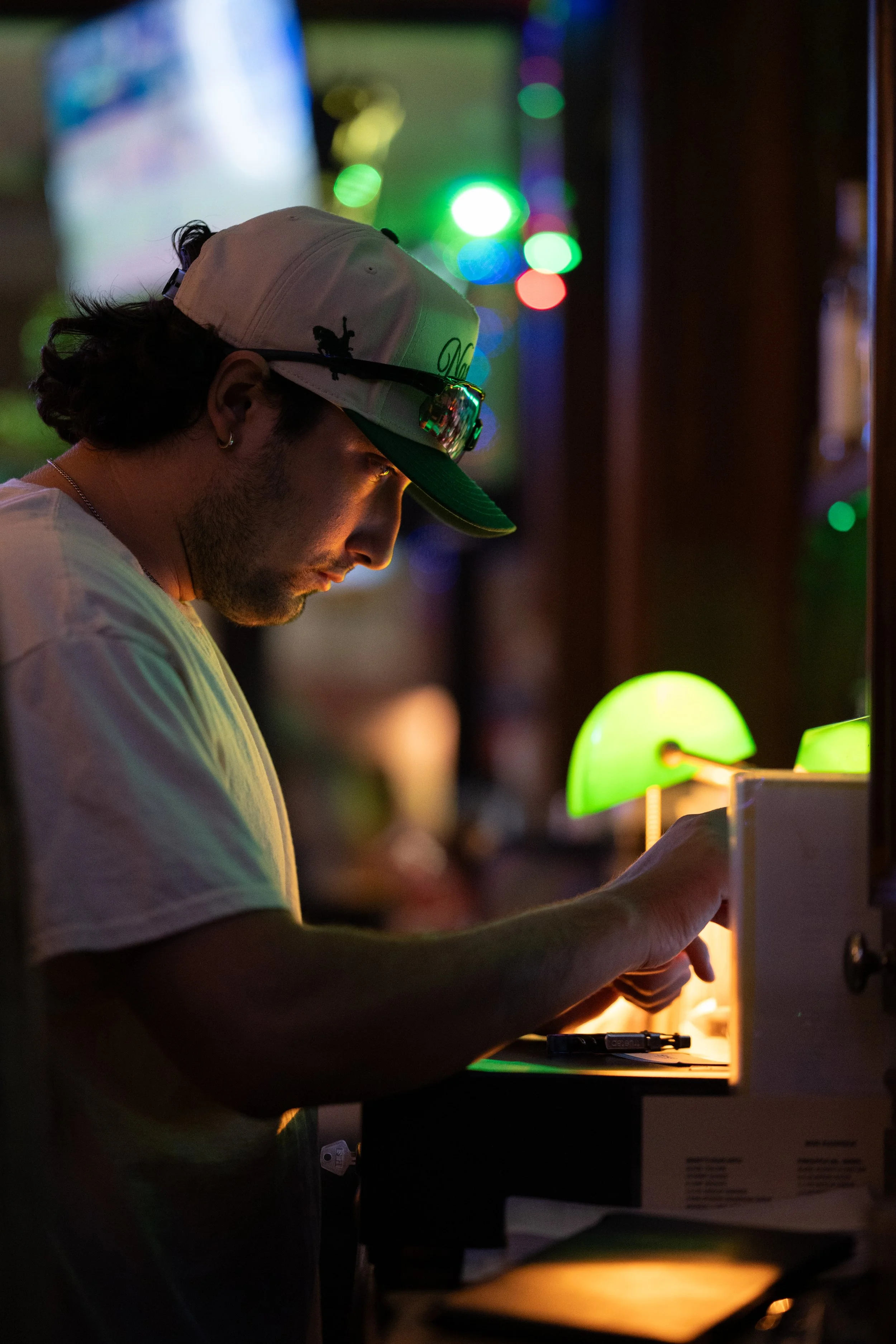 A man wearing a cap with sunglasses on it, working at a counter with a small green lamp, in a dimly lit environment with colorful blurry lights in the background.