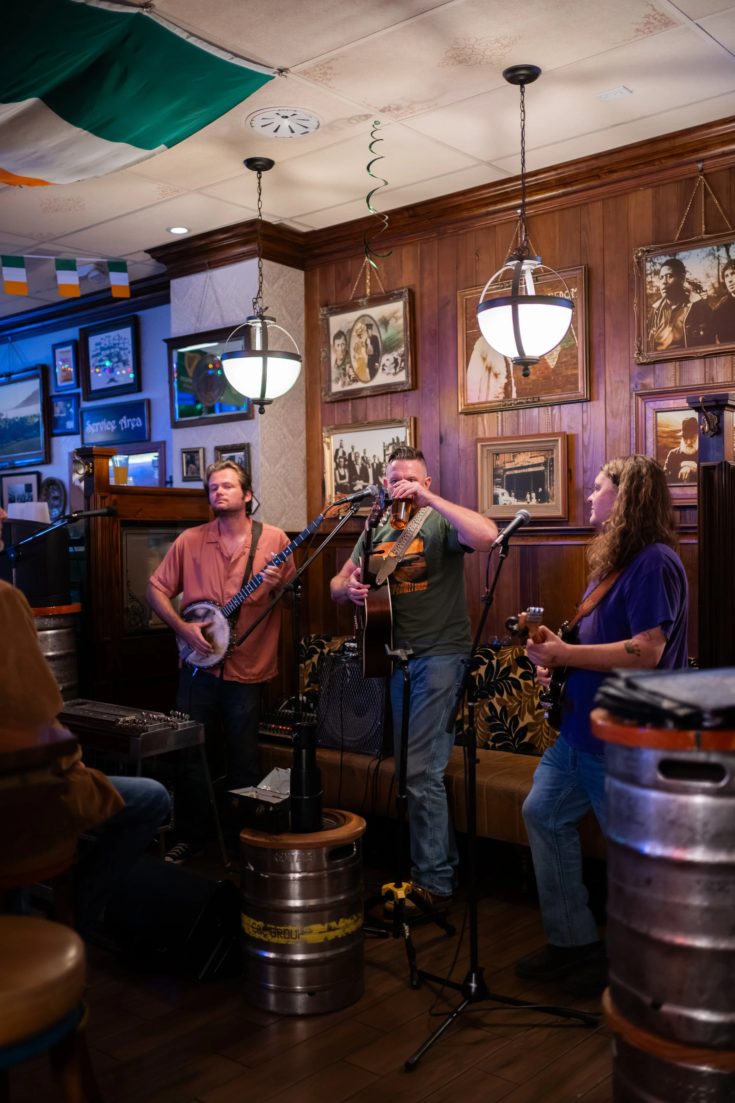 Three musicians perform in a cozy pub decorated with framed pictures on wooden-paneled walls, hanging light fixtures, and Irish flags, with beer barrels used as tables.
