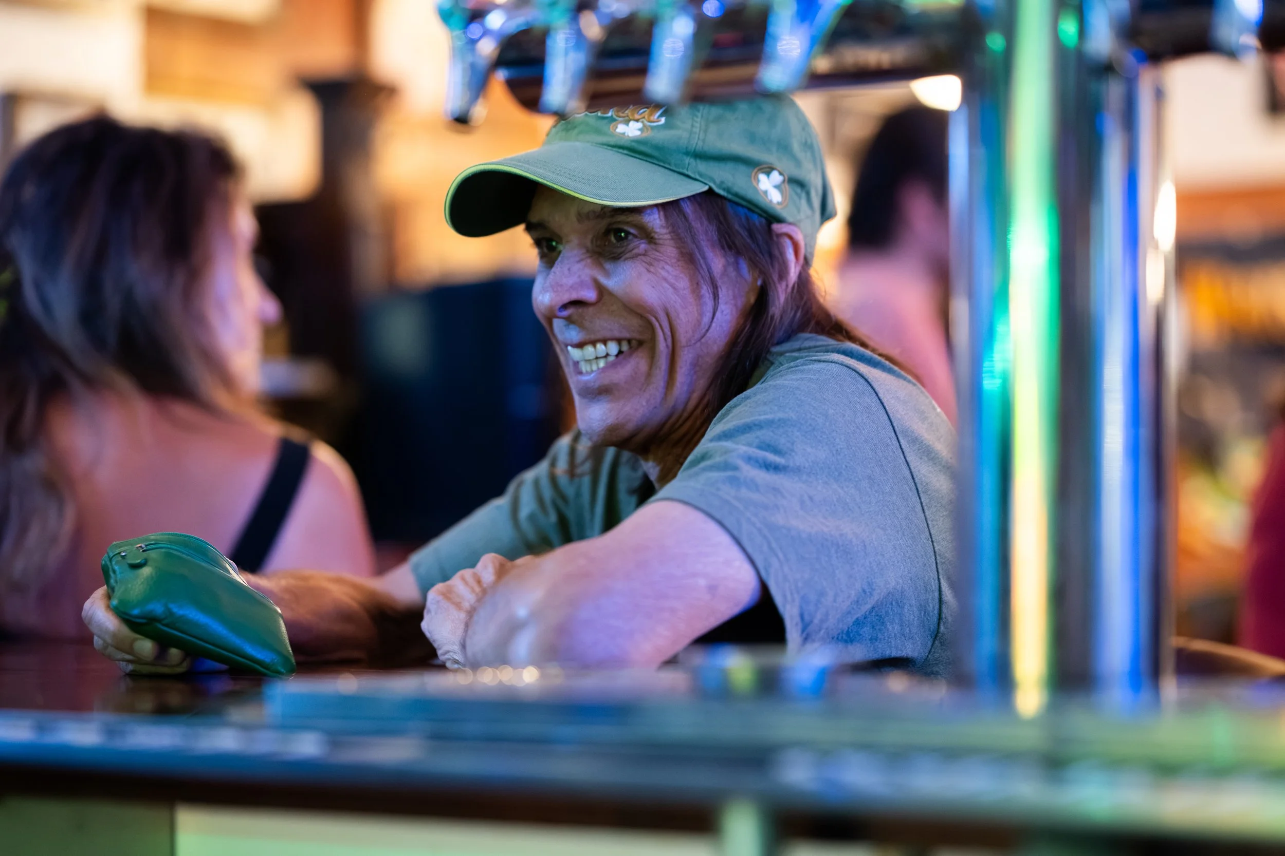 A man with long hair wearing a green cap and a gray shirt, smiling and talking to a woman with brown hair at a bar or pub. They are seated at a dark counter, with colorful neon lights reflecting in the background.