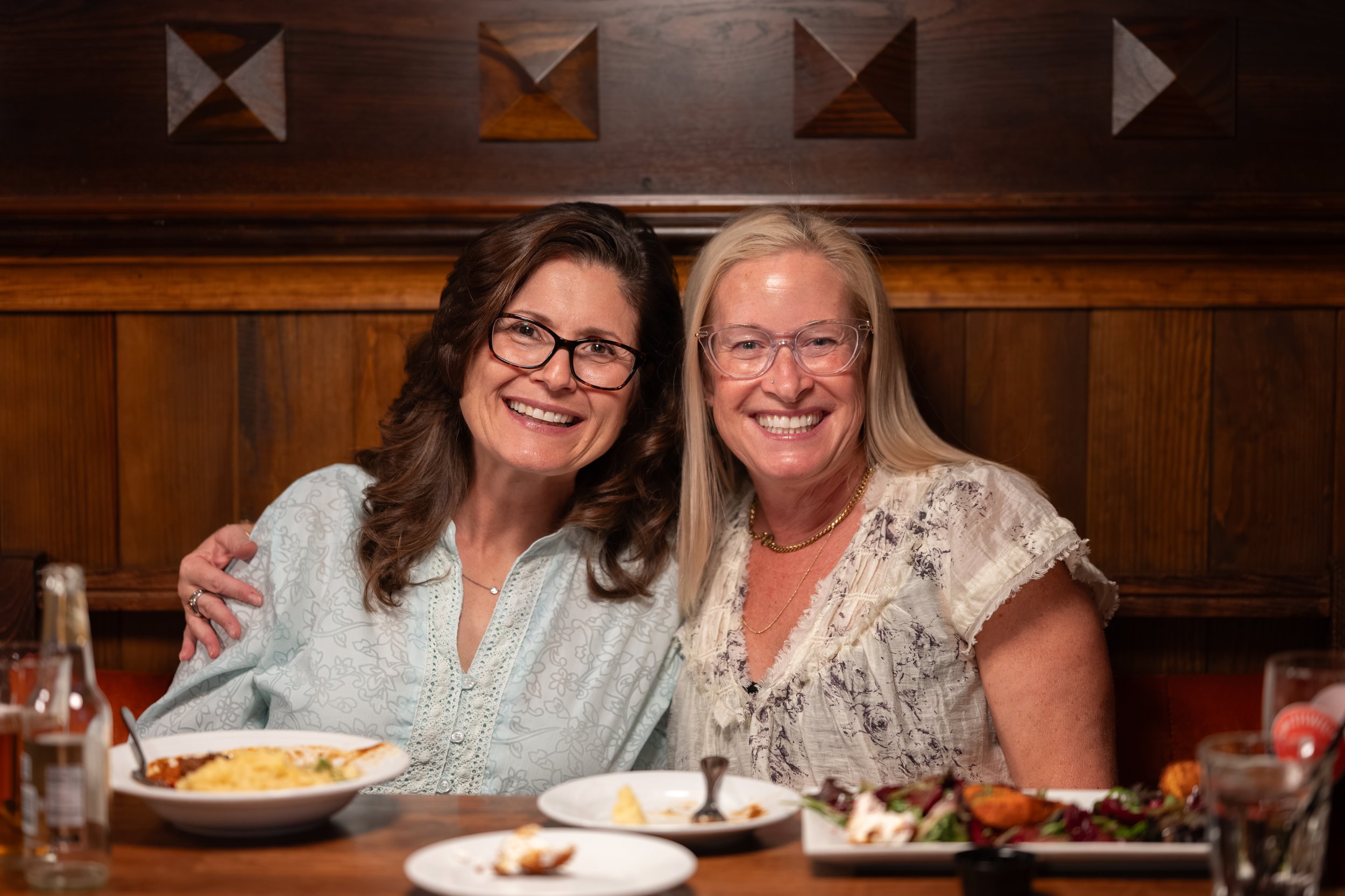 Two smiling women sitting together at a dinner table in a restaurant, with food and drinks in front of them and a wooden wall in the background.