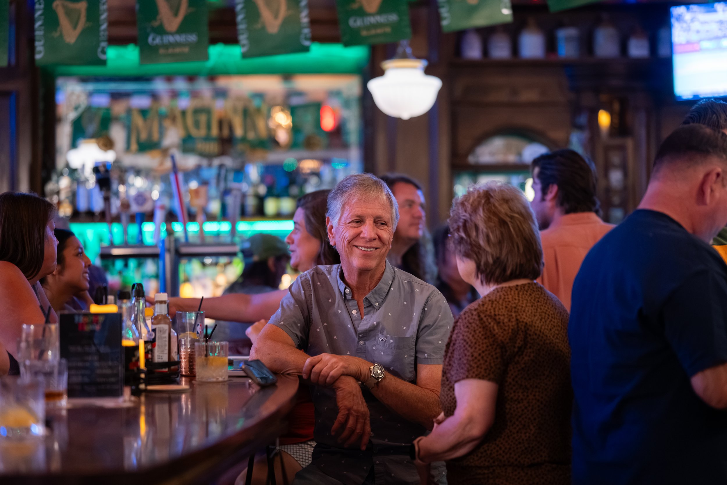 A lively bar or pub scene with a group of people socializing, including a smiling man in the center wearing a light gray shirt, surrounded by others engaged in conversation, with neon lights, banners, and bottles visible in the background.