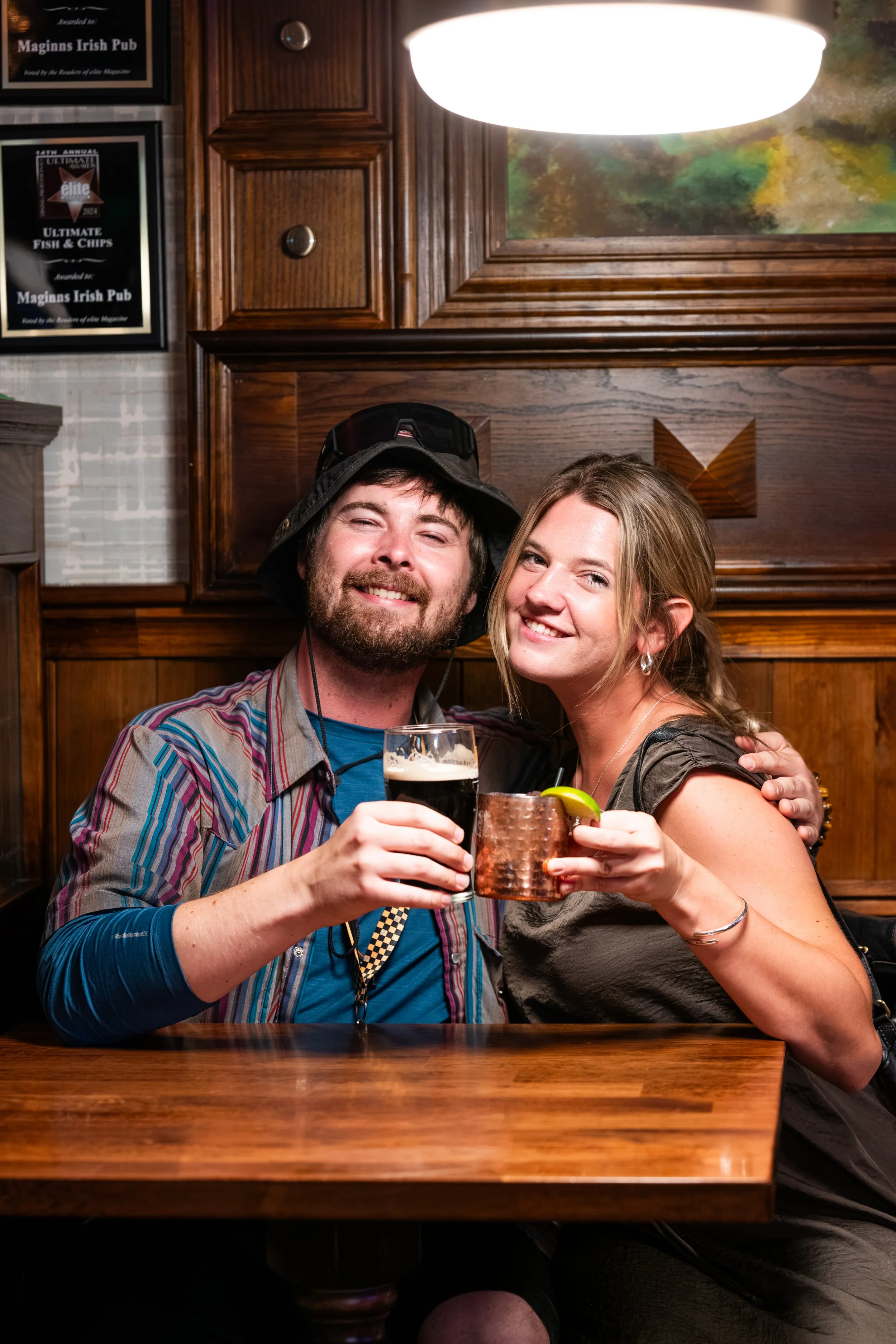 A smiling man and woman sitting closely together in a bar, holding drinks and toasting. The man is wearing a multi-colored striped shirt and a hat, and the woman is in a dark-colored top with earrings. Wooden paneling and restaurant decor are in the 