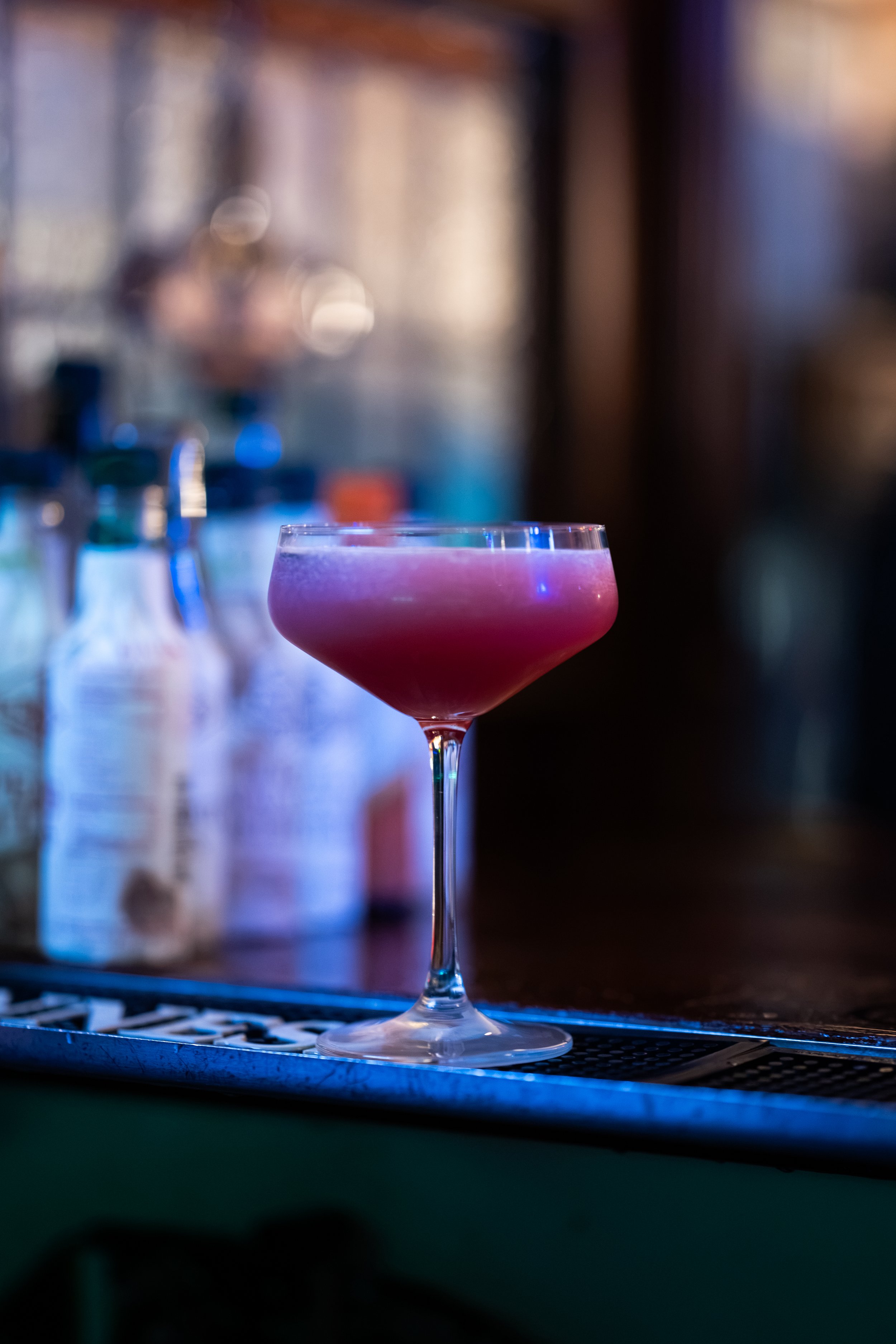 Close-up of a pink cocktail in a coupe glass on a bar counter with blurred bottles in the background.