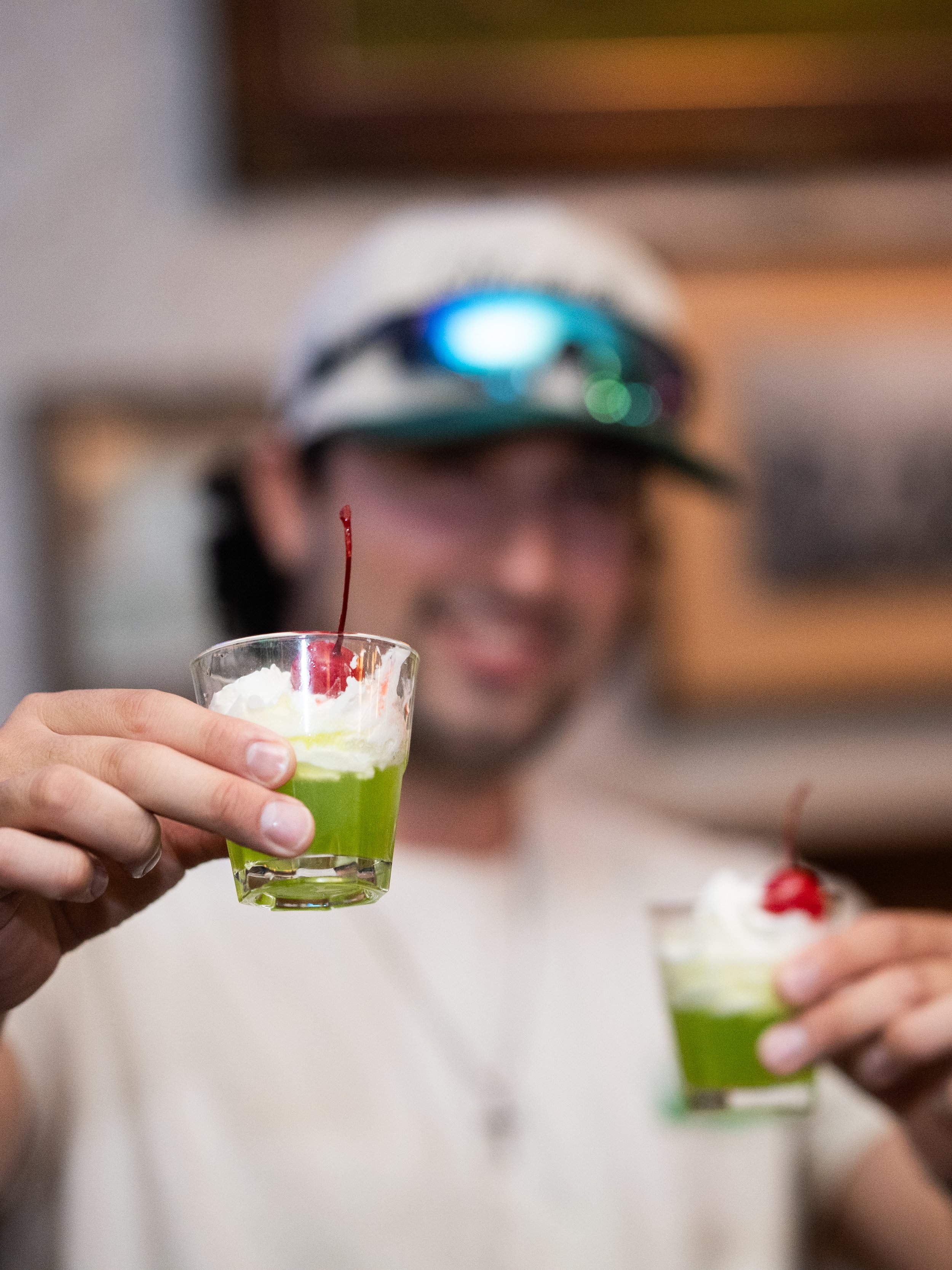 A smiling man holding two small glasses with green desserts topped with whipped cream and cherries, one glass is more in focus.