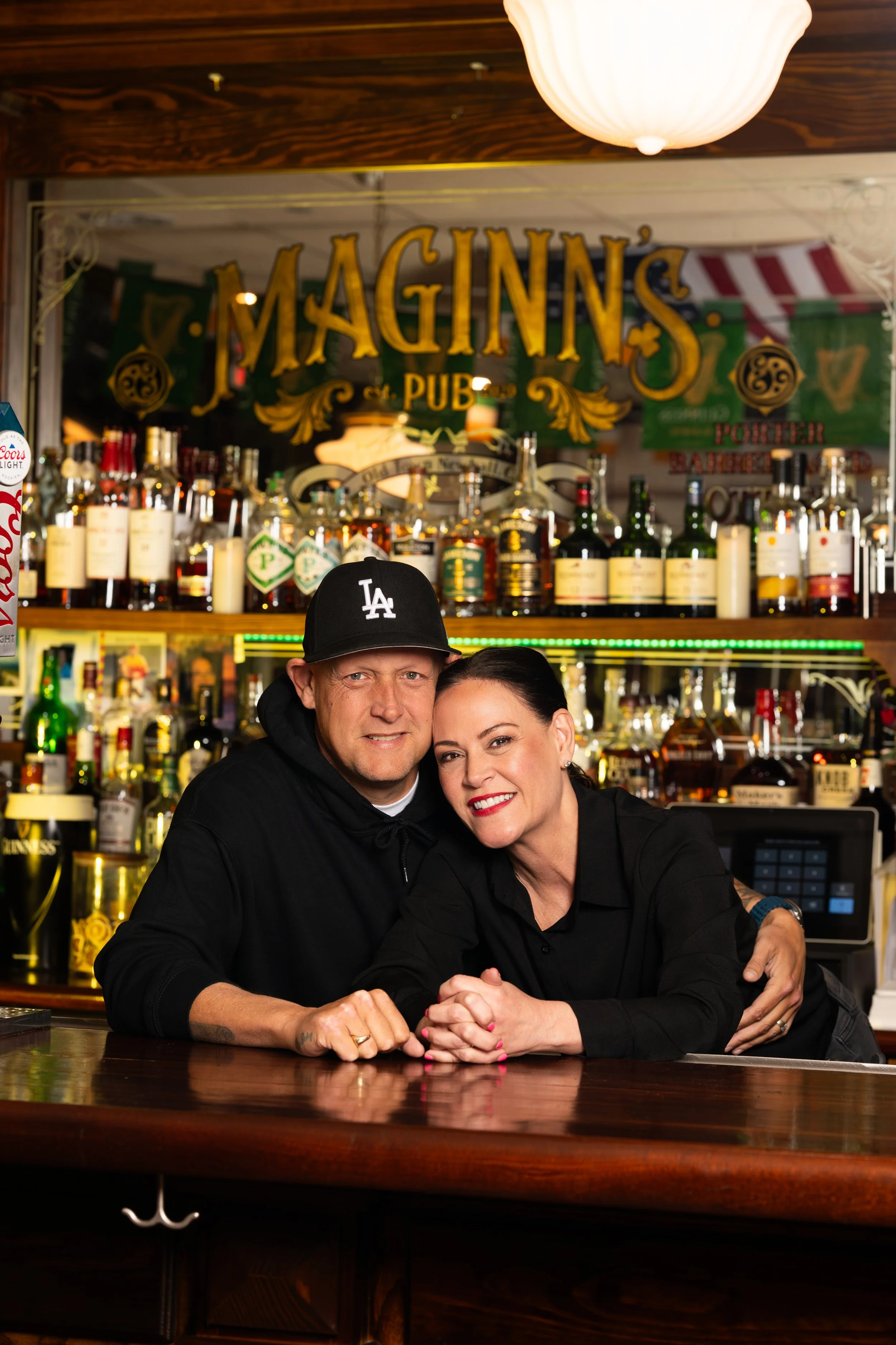 A man and woman smiling and leaning on the bar counter at Maginn's Pub, with bottles of alcohol on shelves behind them.