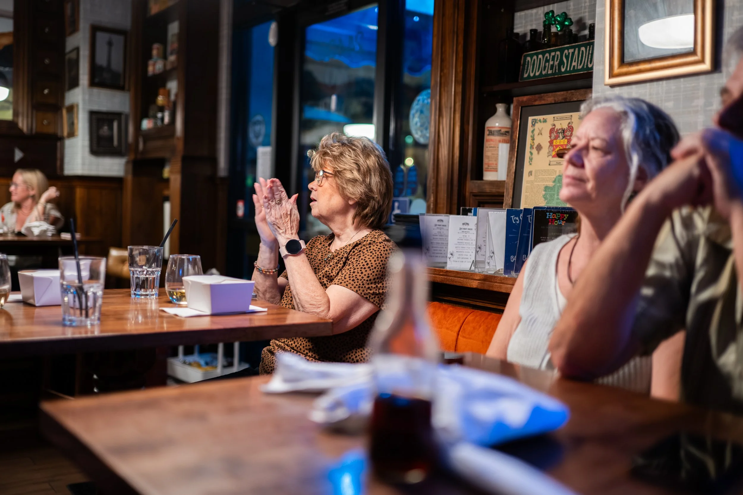Several elderly women sitting at a wooden table in a cozy restaurant, with one woman clapping her hands. The background features wooden shelves with decor and a window showing a dark sky outside.