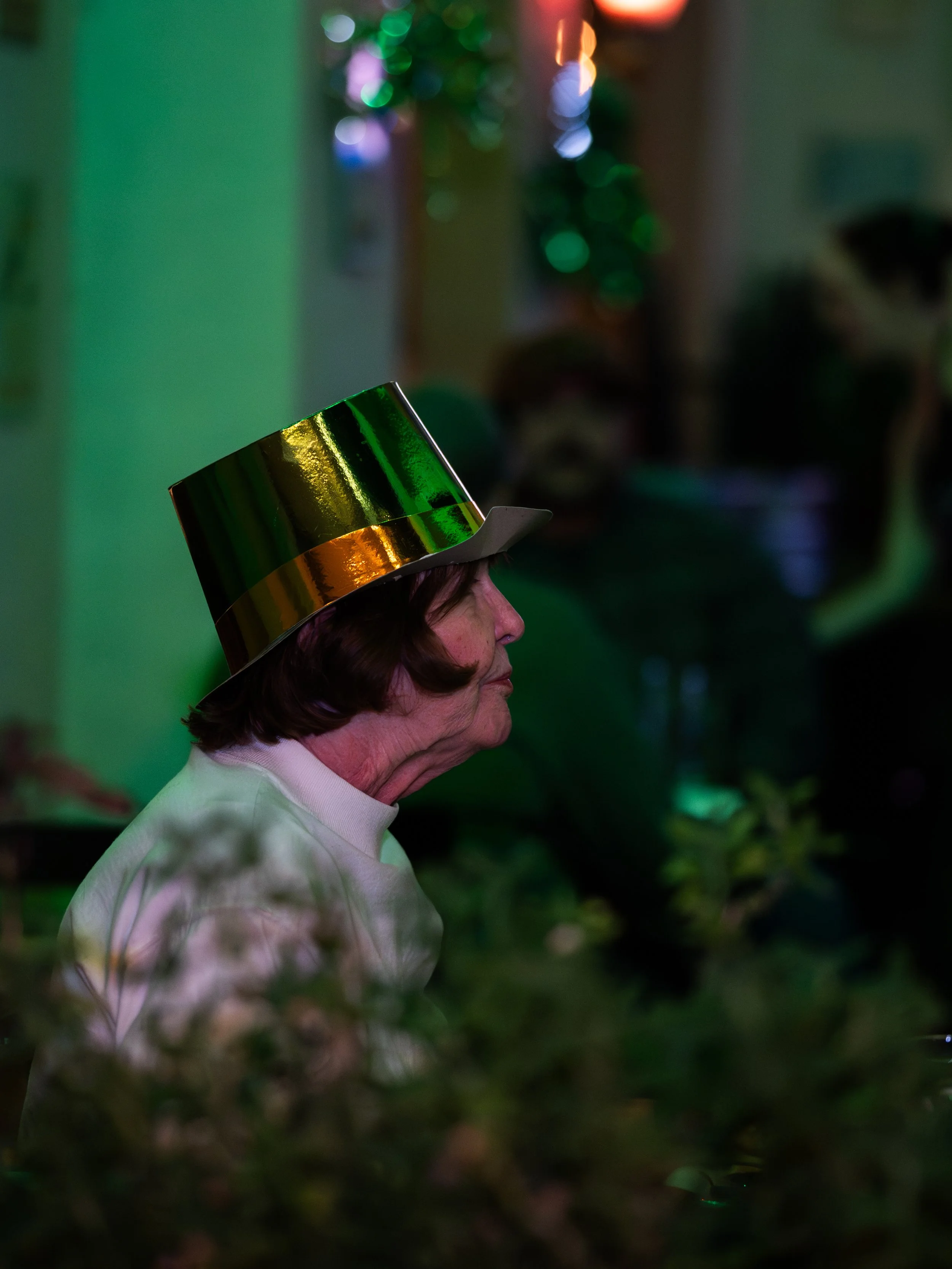 An elderly woman wearing a shiny green and gold top hat sits in profile in a dimly lit room decorated with colorful lights.