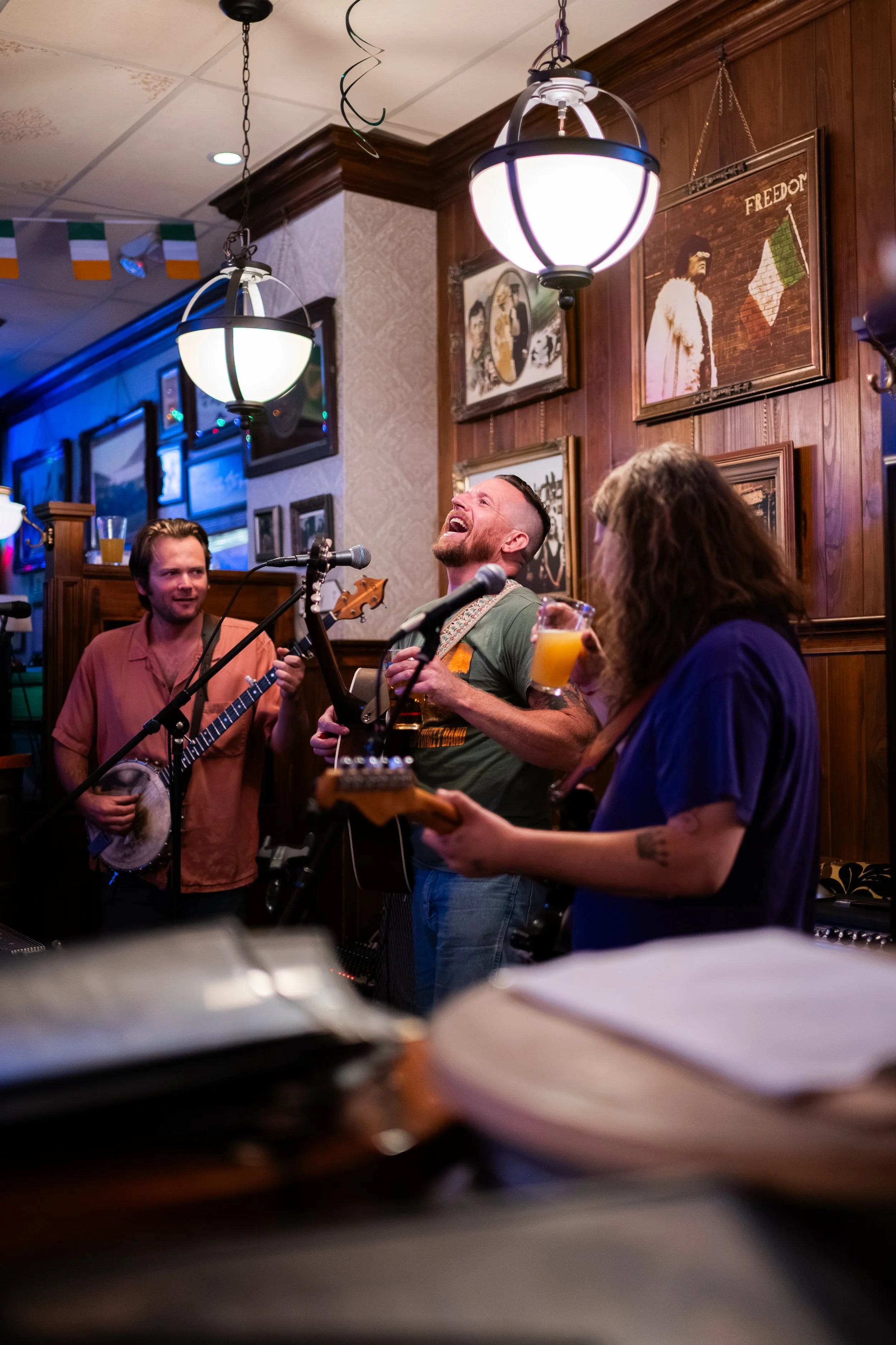 A group of people singing and playing instruments in a restaurant or bar, with framed pictures on wooden walls and hanging lamps.