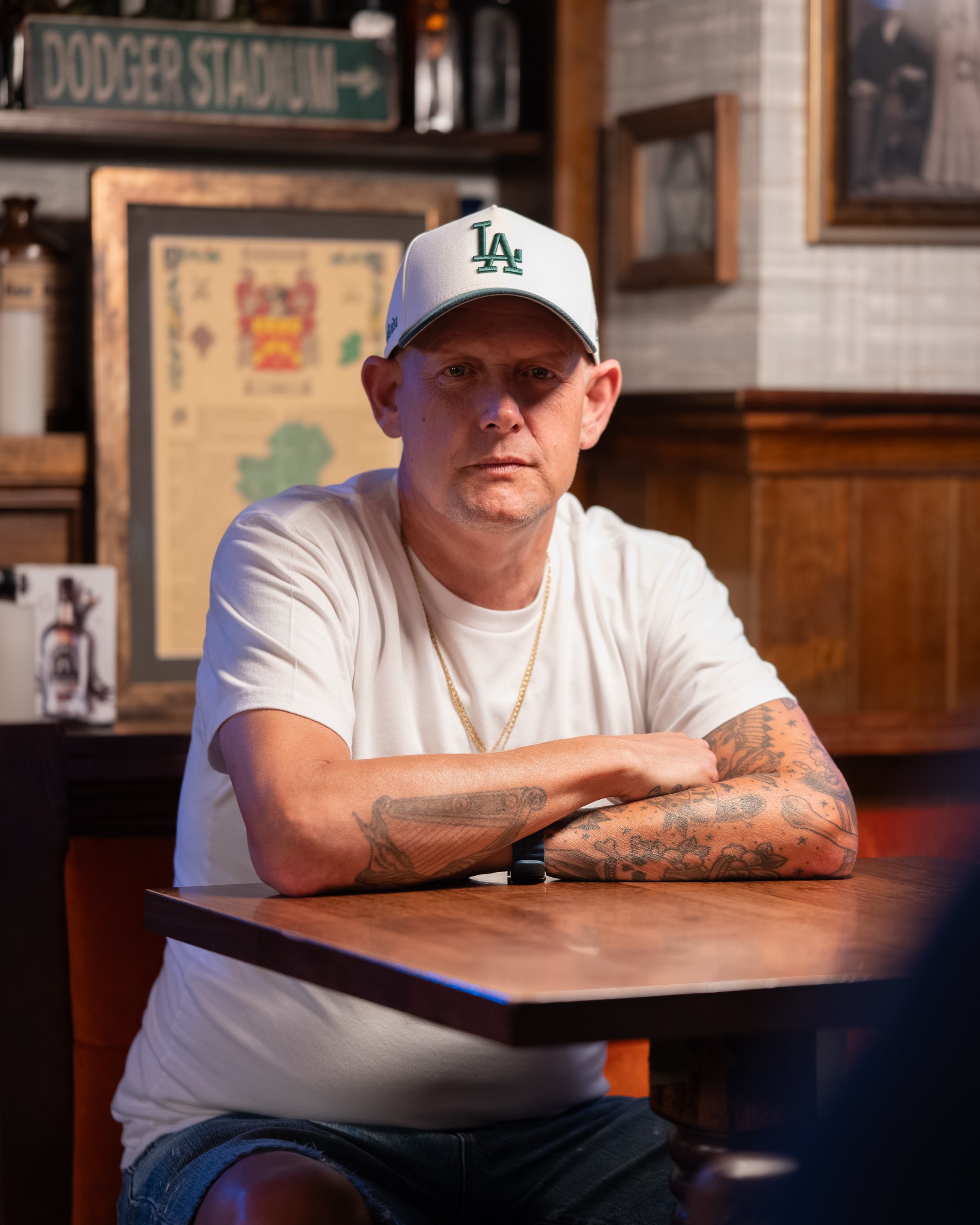 A man in a white LA baseball cap sits at a wooden table with his arms crossed, wearing a white t-shirt and dark shorts, with tattoos on his arms. The background includes framed pictures and a green sign that reads 'DODGER STADIUM' in a rustic, indoor