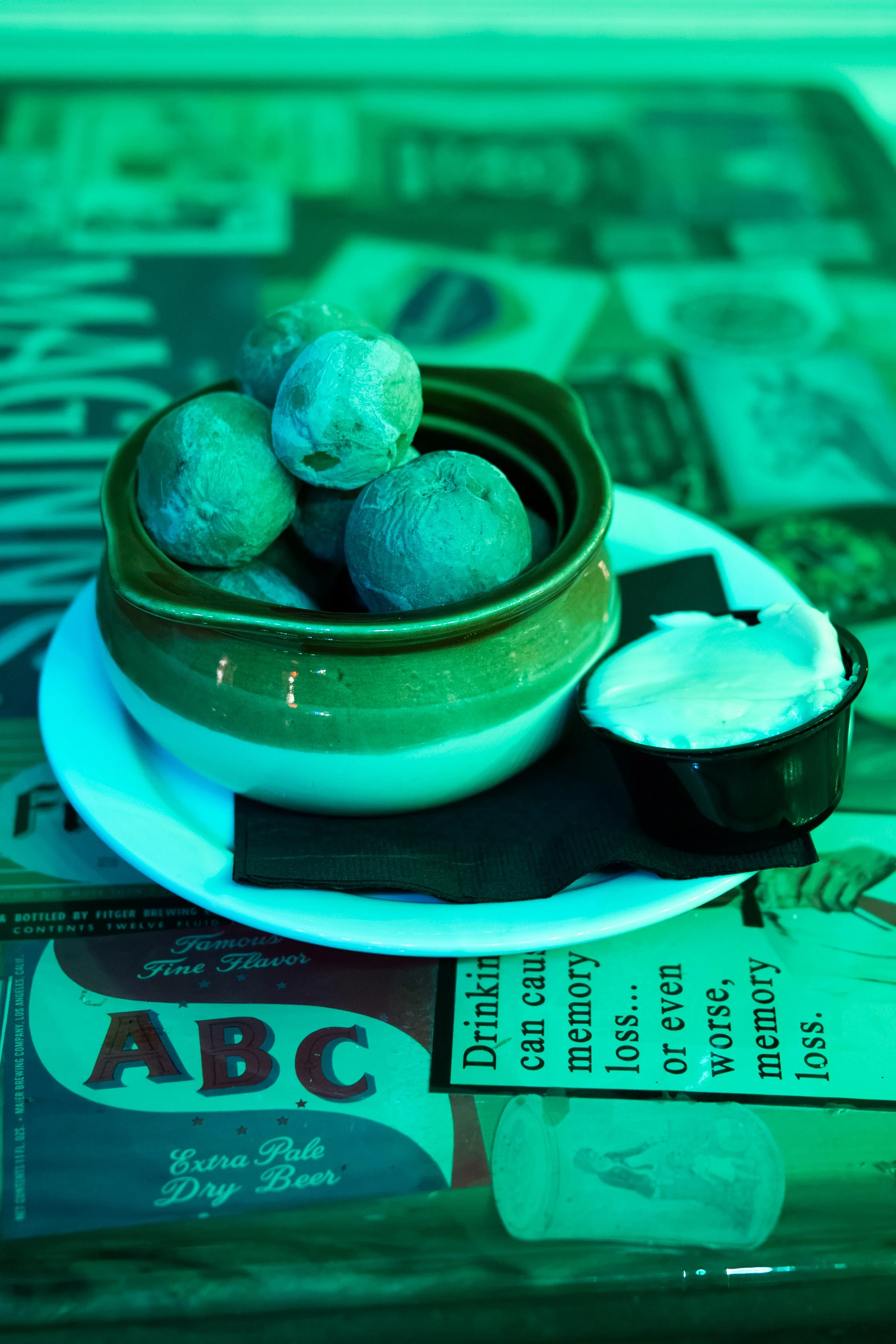 A small green ceramic bowl filled with peeled citrus fruits, possibly limes or lemons, placed on a white plate. There is a small container of white dipping sauce or cream beside the bowl, and the background includes a menu or newspaper with text and 