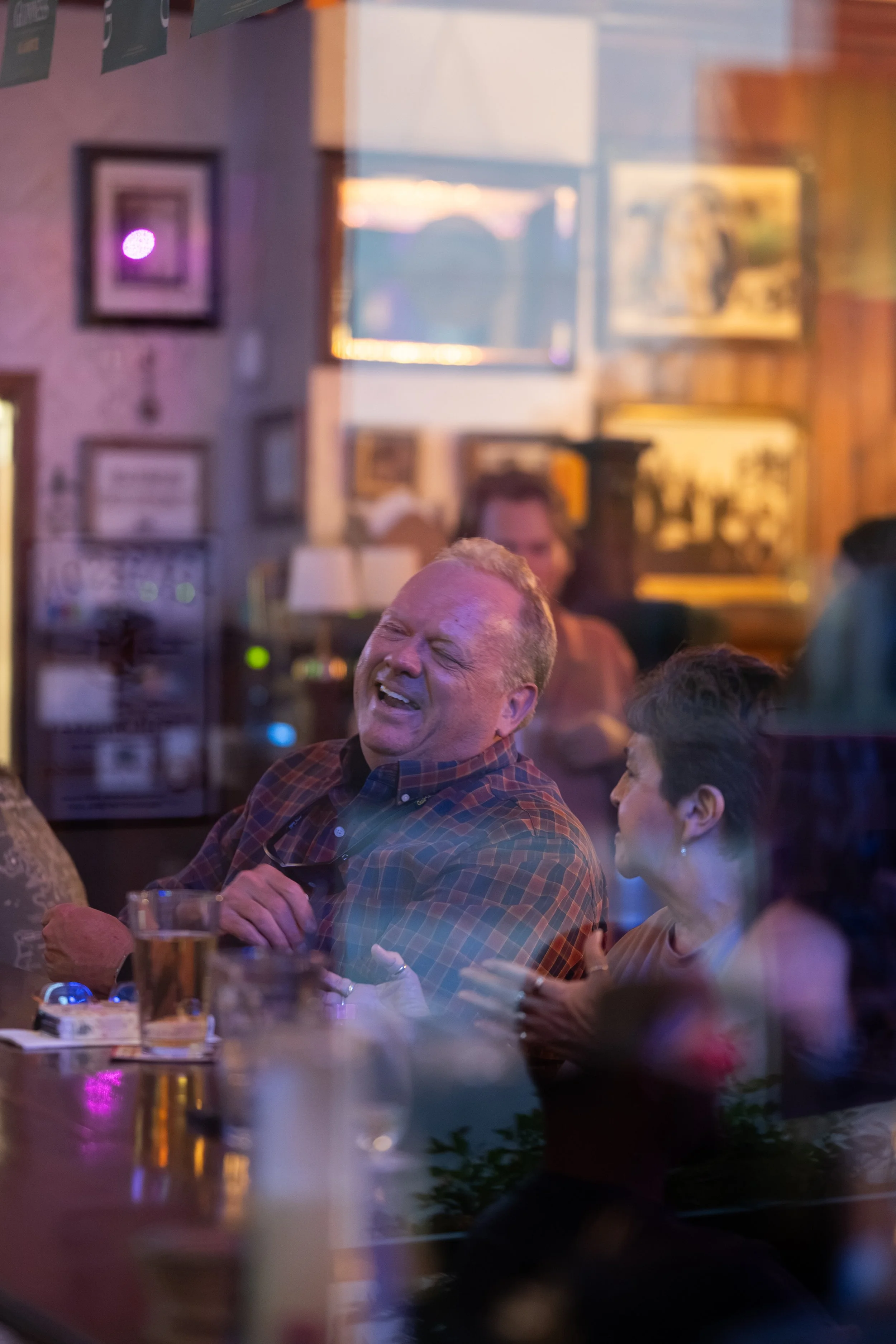 A man with a plaid shirt laughing and holding a glass mug of beer seated at a bar or restaurant with other people, framed pictures, and a window showing a sunset in the background.