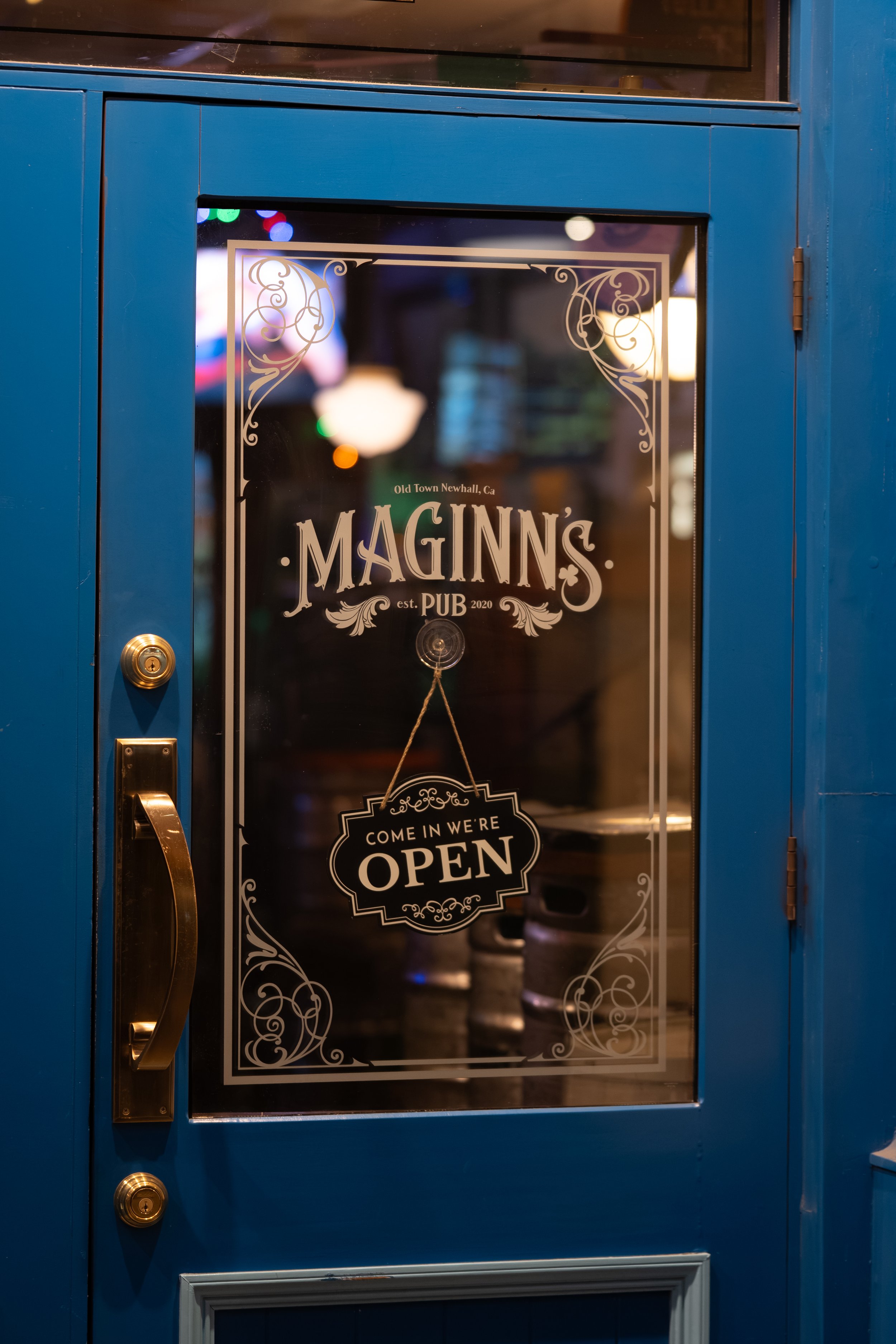 Blue door with a glass window displaying a sign for Maginn's pub, stating it is open and located in Old Town Newhall, California, established in 2020.
