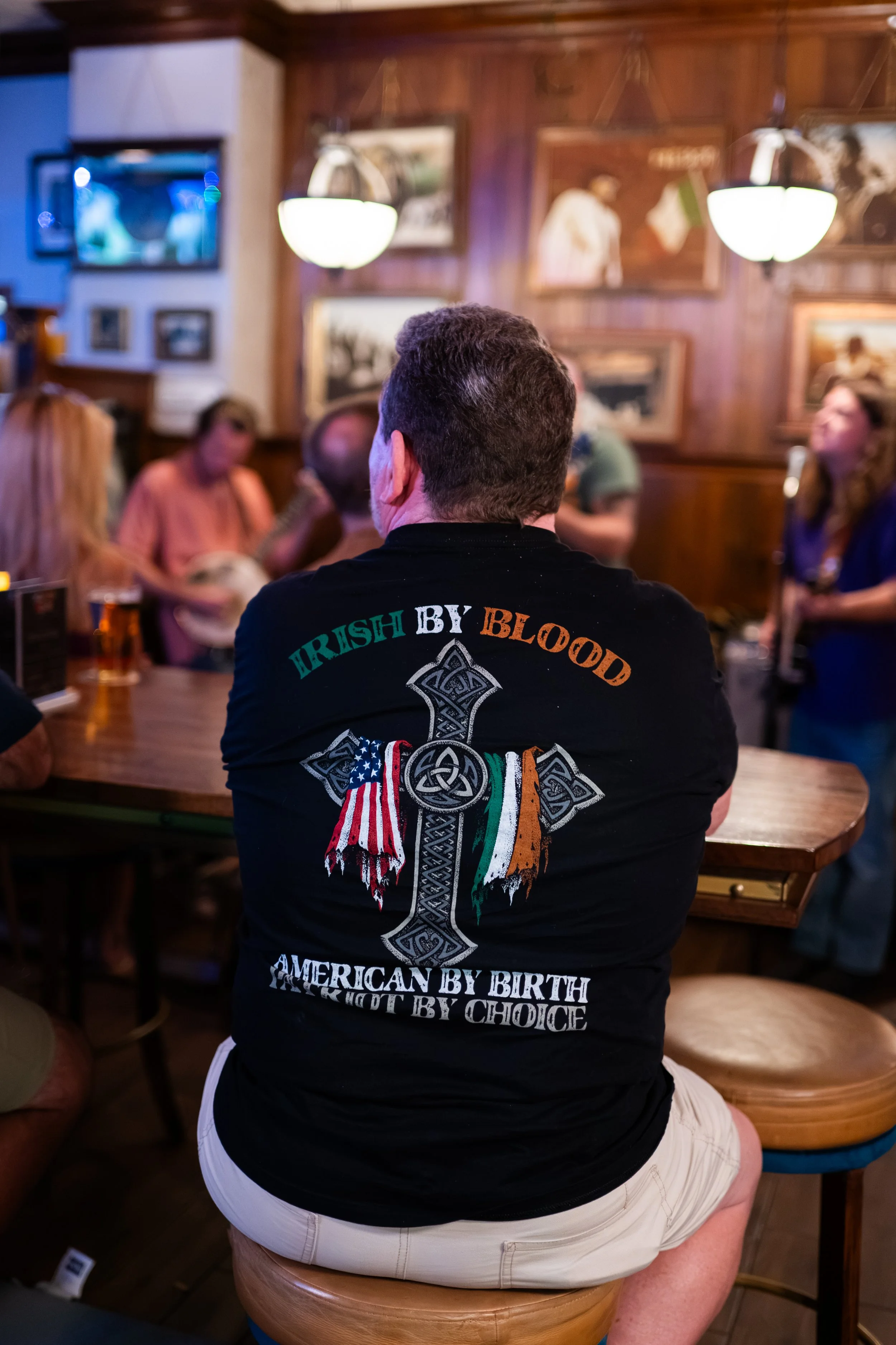 A man with gray hair wearing a black T-shirt with Irish symbols and patriotic flags, and khaki shorts sits on a wooden stool in a dimly lit pub, surrounded by patrons and vintage photos on the wooden walls.