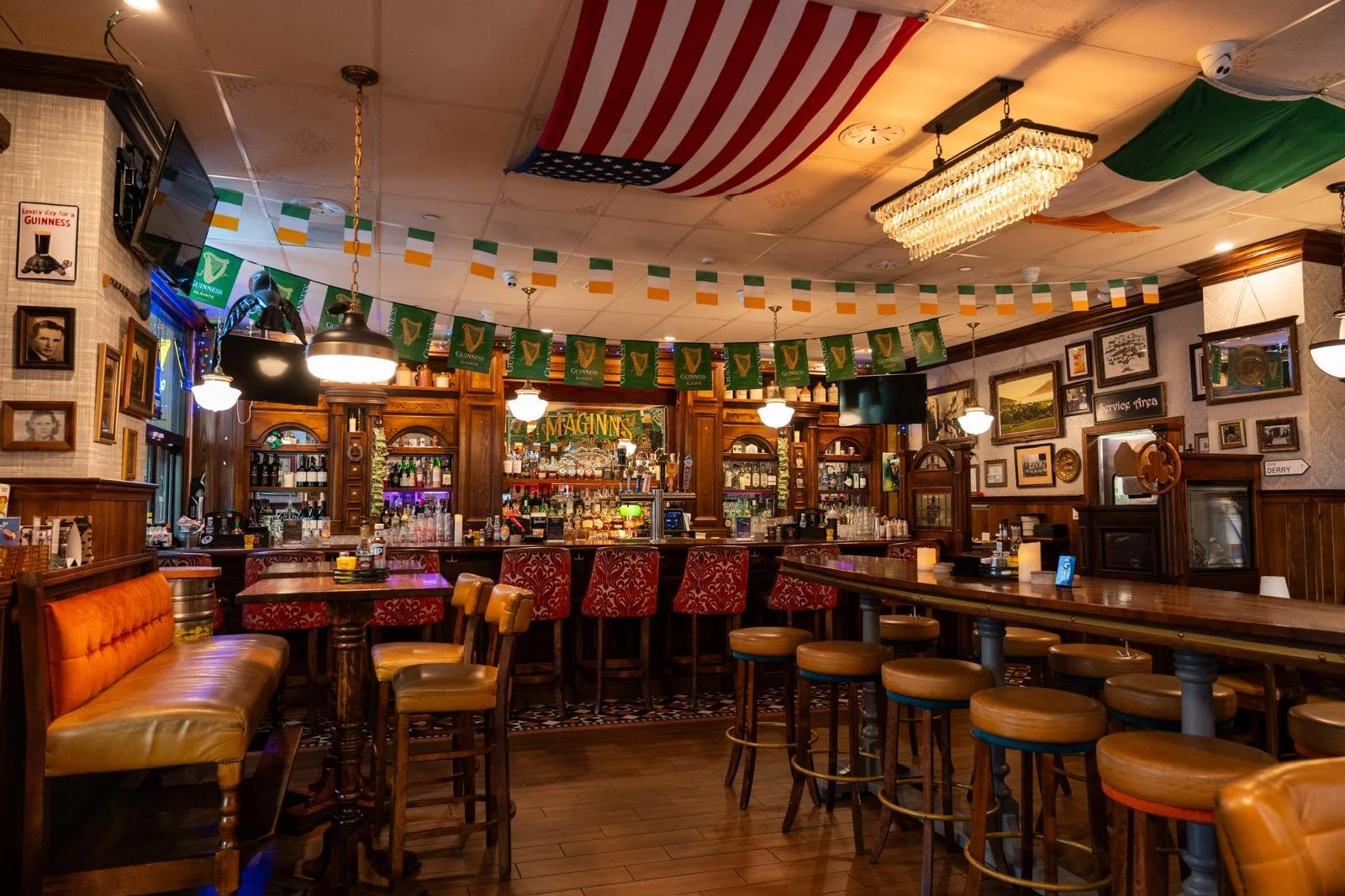 The interior of a pub decorated for St. Patrick's Day with Irish flags and Guinness banners, featuring a bar area with stools, framed pictures, and festive decor.