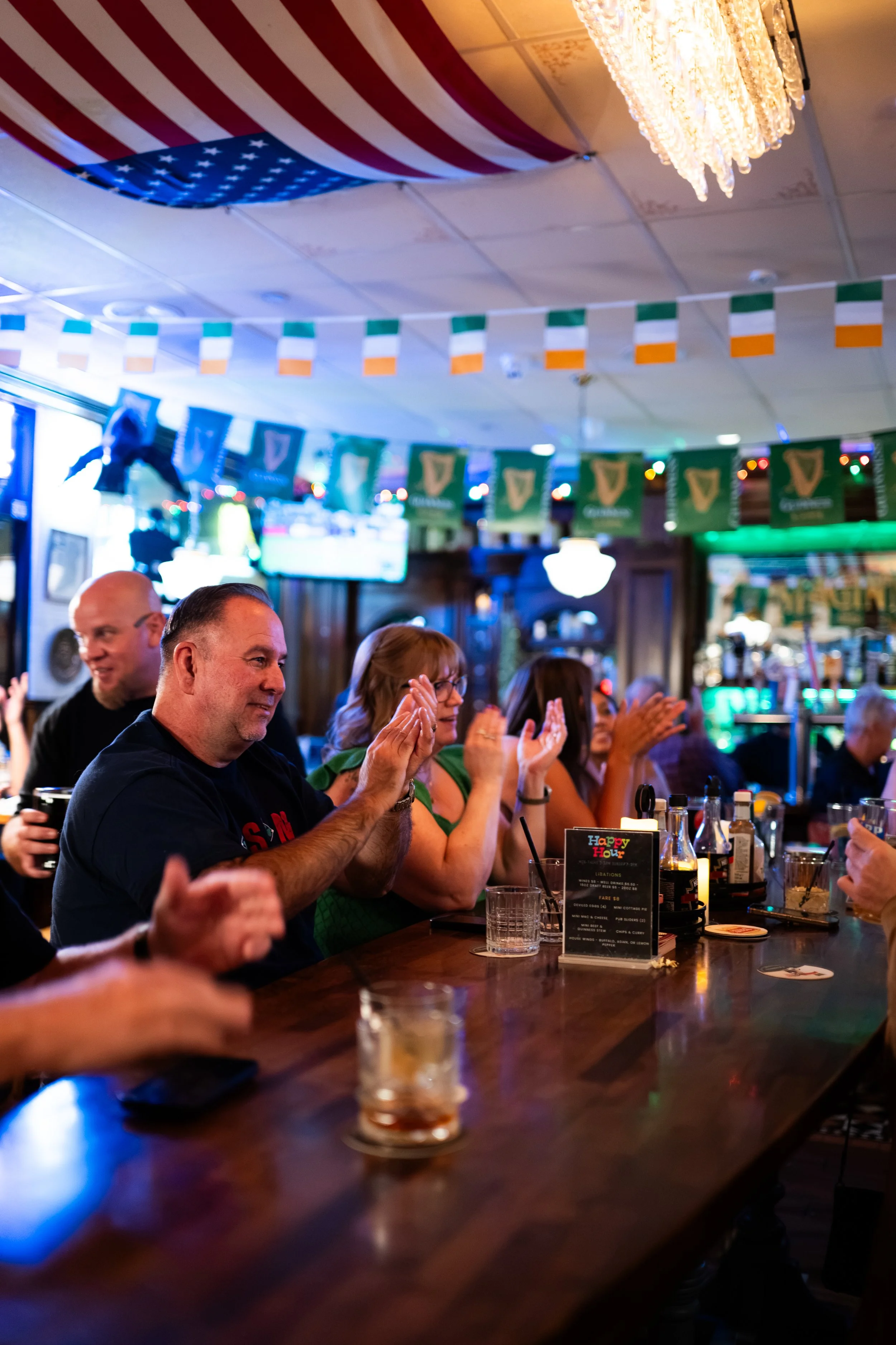 People sitting at a bar with an American flag decoration on the ceiling, some of them clapping and smiling, and a menu on the bar with the words 'Happy Hour' visible.