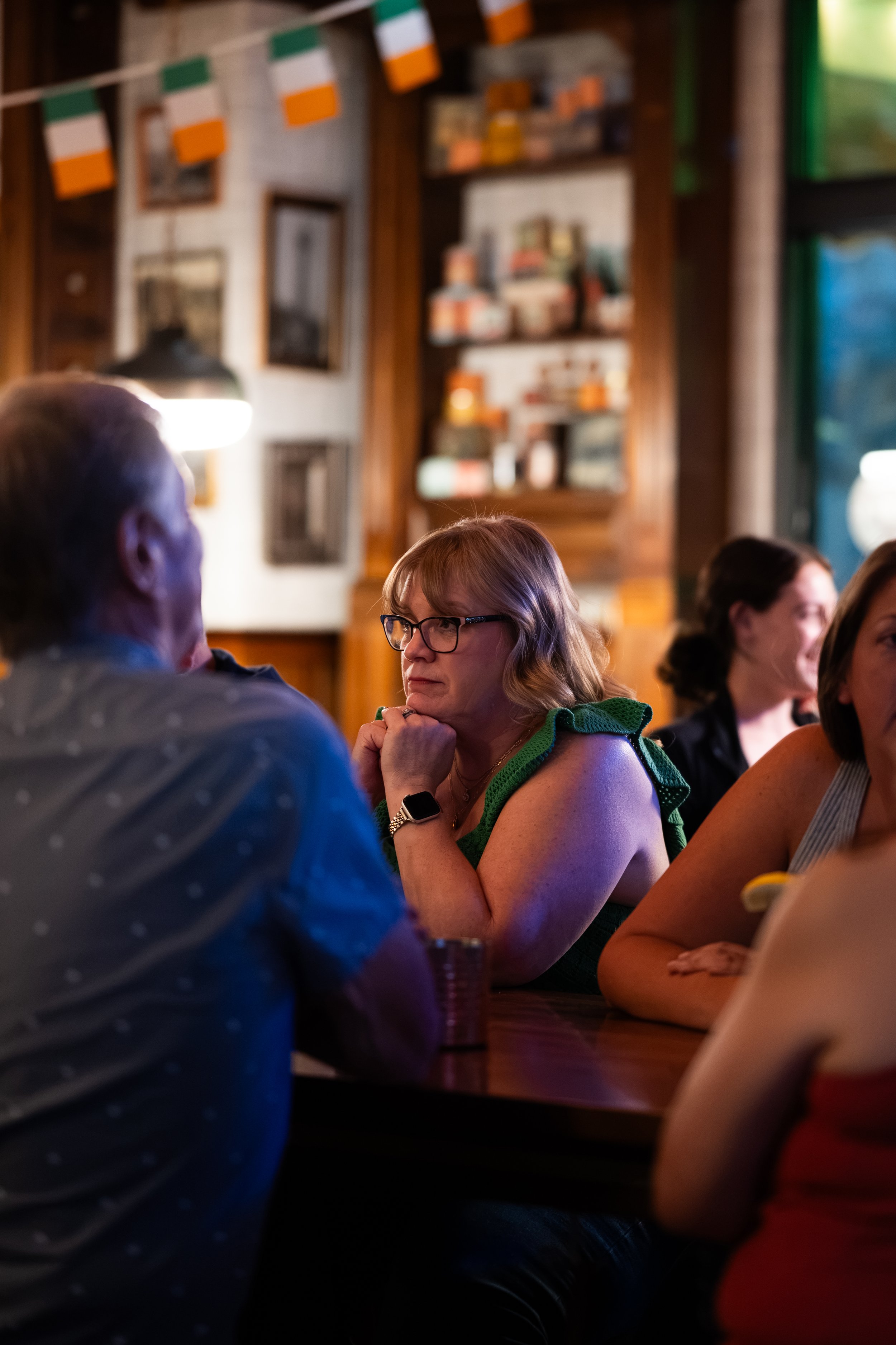 A woman with glasses and a green sleeveless top sitting at a bar or restaurant table, resting her chin on her hand, with other people seated nearby. The background shows wood-paneled walls, shelves with decorative items, and Irish flags hanging.