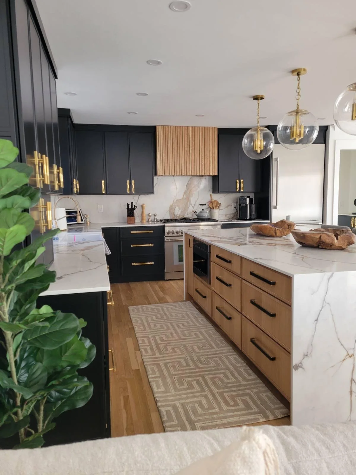 Modern kitchen with black upper cabinets, light wood lower cabinets, marble countertops, and a marble island. Includes pendant lights, a stainless steel refrigerator, stovetop, coffee maker, and decorative wooden bowls. Hardwood floors and a beige ge